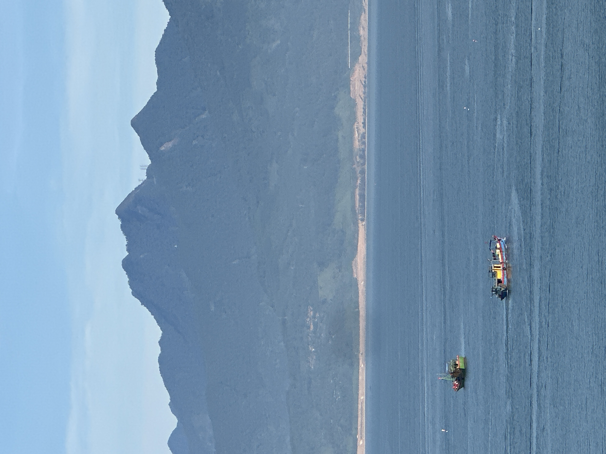 The image depicts a wide shot of the sea with mountains in the background and two fishing boats on the water. The sea is a deep blue, with ripples indicating a gentle movement. The mountains are a muted blue-grey, suggesting they are distant. The sky is a pale blue with some wispy clouds. One fishing boat, predominantly green, is positioned in the upper left quadrant of the image, and another, brightly colored with yellow, red, and blue accents, is in the lower right quadrant. The boats appear to be moving. The shoreline is visible between the sea and the mountains, with some lighter colored sand or land. This scene is characteristic of the coastline near Đà Nẵng, Vietnam, likely facing the South China Sea, with the Sơn Trà Peninsula or the Hải Vân mountains visible in the distance.