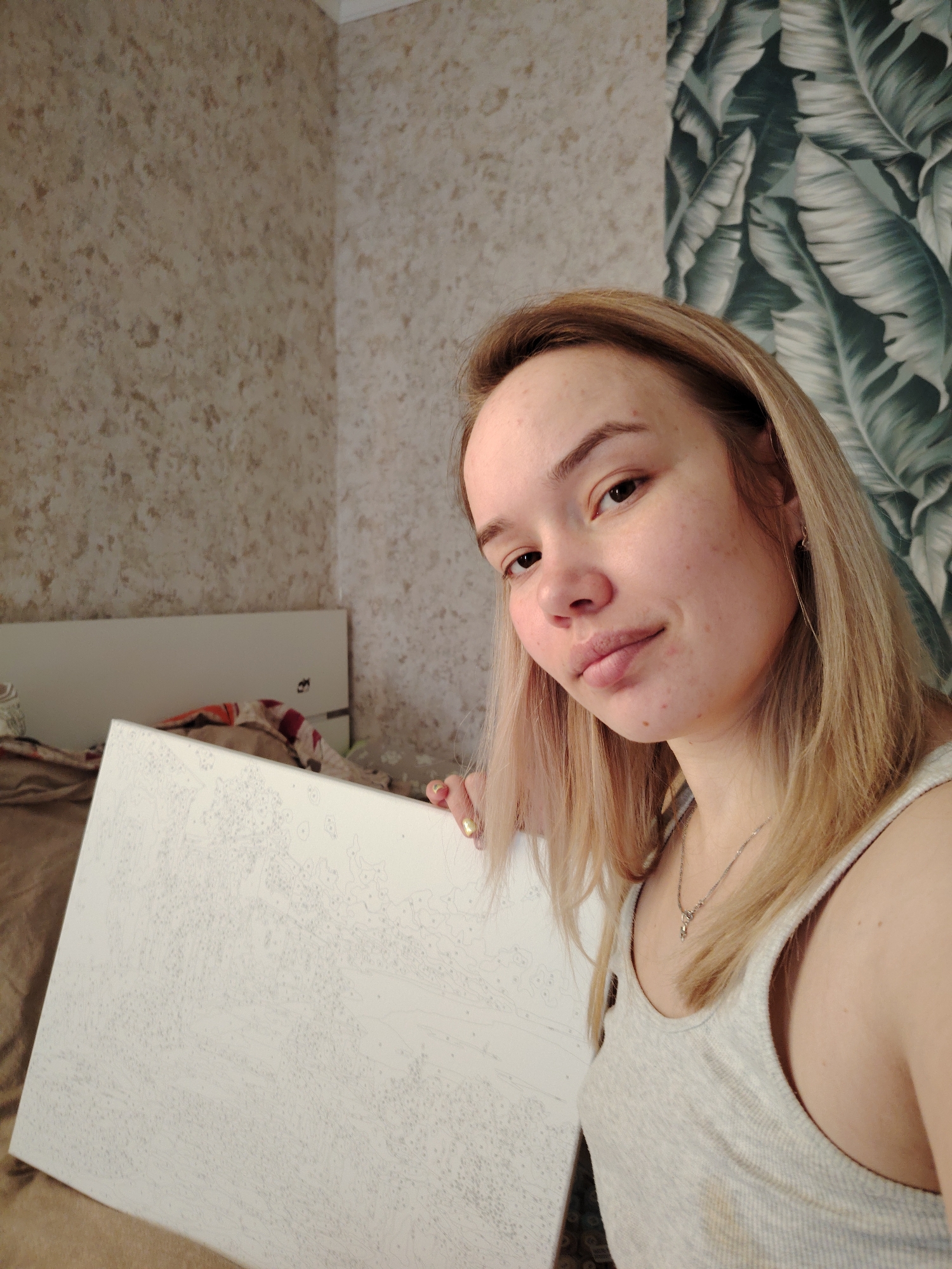 The photograph shows a young woman in a room in Марковский, Russia. She is fair-skinned with blonde hair, wearing a light grey tank top and a silver necklace. She is holding a large, white canvas with a paint-by-numbers outline. Behind her is a bed with a headboard, covered in bedding. The wall has two different wallpapers: one with a beige, textured pattern and another with a large, green leaf print. The lighting in the room is soft and natural.