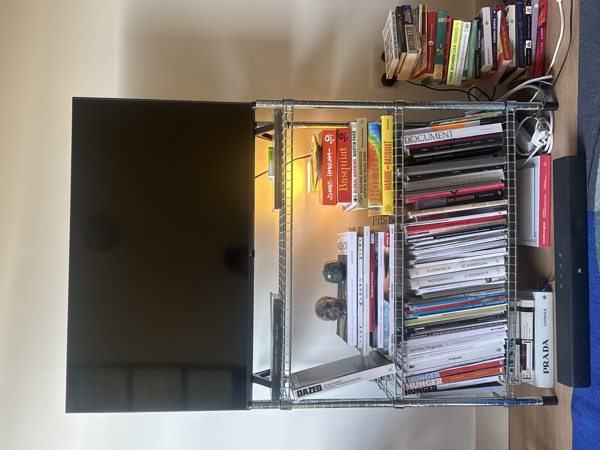 The image shows a shelf unit with a television on top, a stack of books to the right, and a soundbar on the floor below. The shelf unit is silver metal with wire shelves. On the top shelf, there is a single book. The second shelf from the top contains a book titled "DAZED" and a stack of several other books, some with colorful covers. On this shelf, there are also two decorative objects, one resembling a skull and the other a small green orb. The third shelf from the top is filled with many magazines and books arranged vertically. The bottom shelf has a book titled "PRADA CATWALK" and another book with red and white text. Wires and electrical plugs are visible on the floor behind the shelf unit. To the right of the shelf unit, a tall stack of books leans against the wall. The background is a plain wall. No people are visible in the image. The environment appears to be an indoor living space, possibly a home or an office. There is no information in the image to indicate the location in the city of London, United Kingdom.