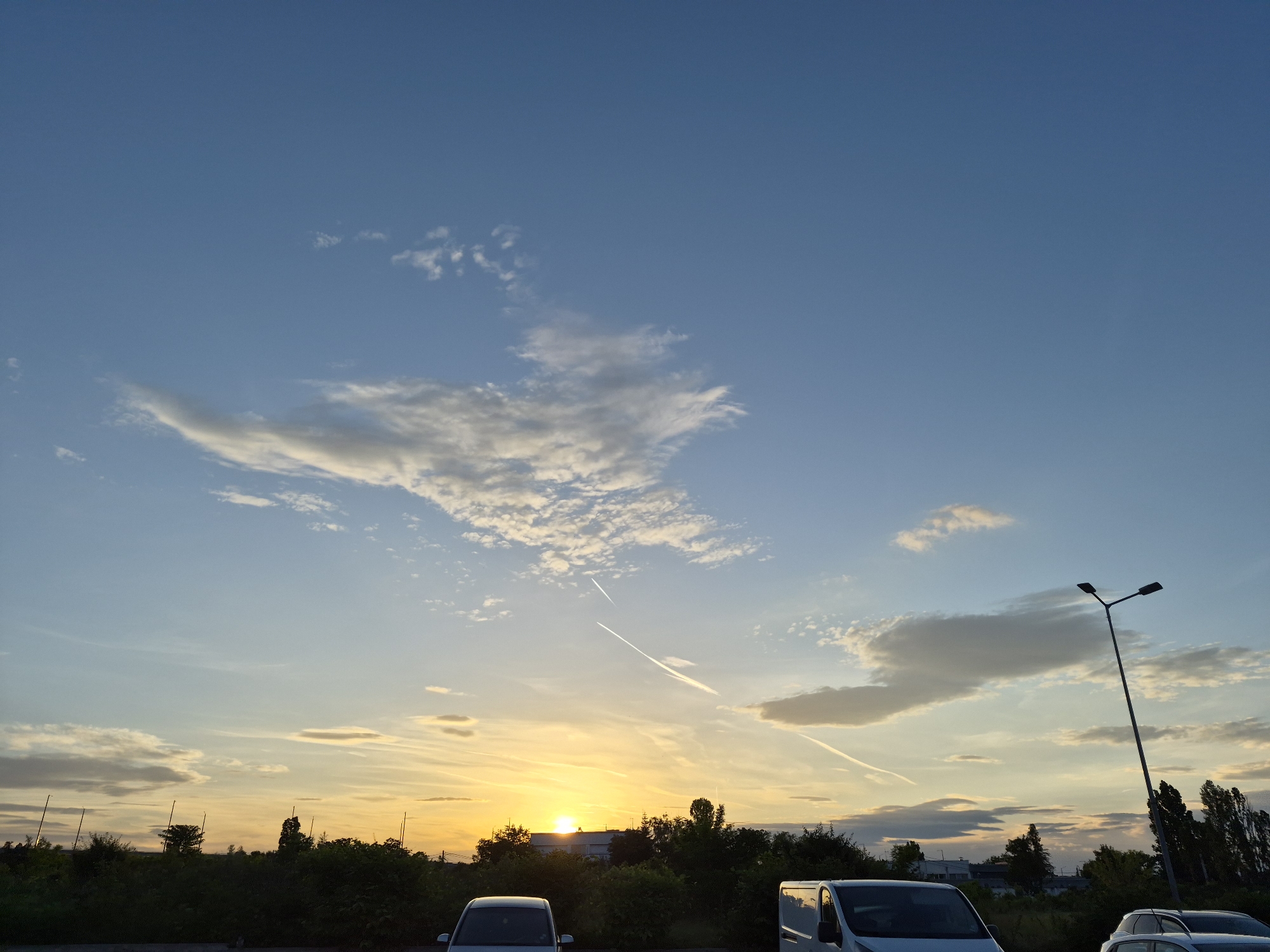 The image shows a sunset in what appears to be a parking lot or roadside area in Sofia, Bulgaria. The sky is primarily blue with scattered clouds, including some contrails from aircraft. The sun is setting near the horizon, creating an orange and yellow glow. Below the horizon line are trees and vegetation. Several vehicles, including what appear to be vans and cars, are parked in the foreground. There is also a tall street light pole on the right side of the image. No people are visible.