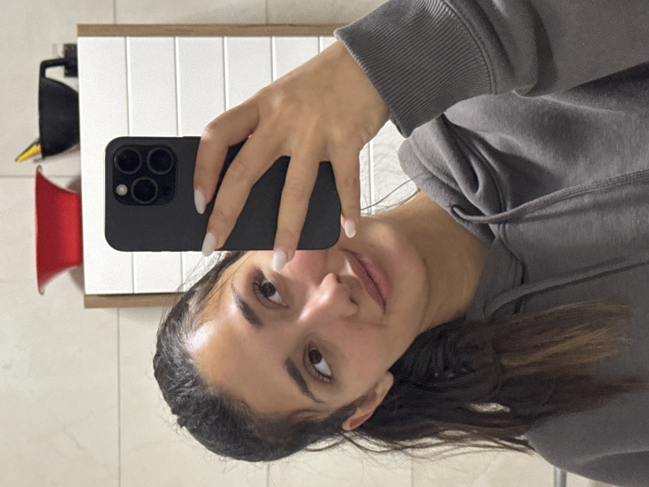 A woman in a grey hoodie takes a selfie in front of a bathroom mirror. Her hand, with manicured light-colored nails, holds a black smartphone with multiple camera lenses. A red object and a black item hang on the tiled wall behind her. The mirror reflects white tiles. Her brown hair is pulled back, and she looks into the camera.