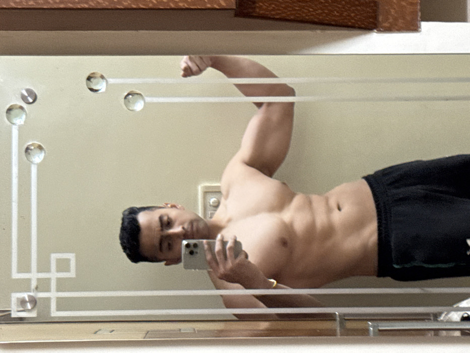 The image depicts an adult male taking a mirror selfie indoors in Kolkata, India. The male is shirtless, revealing a muscular physique with defined chest, abdominal muscles, and biceps. He wears black athletic shorts. In his left hand, he holds a light-colored smartphone vertically, capturing his reflection. His gaze is directed towards the phone screen, and his right arm is bent, flexing the bicep with the fist clenched. A thin gold-colored bracelet is present on his left wrist. The mirror features an etched geometric border with square and line patterns, along with several clear, circular glass embellishments. Behind the subject's reflection, a plain, light-colored wall is visible, on which a white electrical switch plate with a circular switch is mounted. A segment of a brown-toned wooden door frame or paneling is visible on the right side of the reflection.