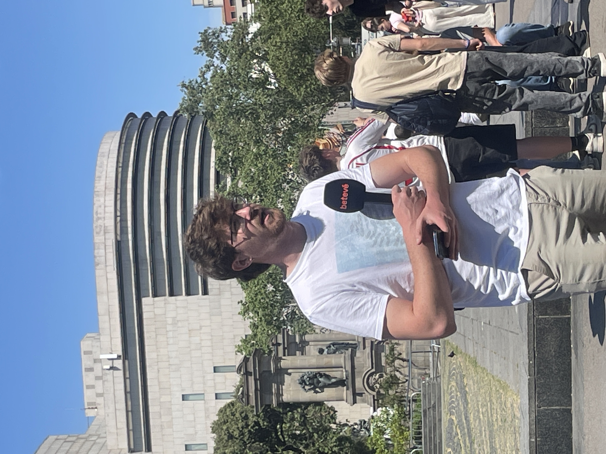 The image depicts an outdoor urban scene on a bright, sunny day, likely in Barcelona, Spain. In the foreground, a man with brown, curly hair and glasses, wearing a white t-shirt and khaki pants, is holding a black microphone in his right hand and a smartphone in his left. The microphone clearly displays the white text "betevé", indicating he is a reporter or interviewer.
Several other individuals, dressed in casual attire such as t-shirts, jeans, and sneakers, are visible in the background, standing or walking. Behind the main subject, a distinctive modern building with a grey, multi-ribbed, curved roofline dominates part of the skyline. Below this curved section, the building's facade is composed of lighter-colored stone blocks with rectangular windows. To the right, a large, ornate stone monument featuring dark sculptural details is present. Lush green trees are interspersed throughout the background, providing natural accents to the urban environment. The clear blue sky suggests good weather, and the strong sunlight casts visible shadows on the paved ground. The scene appears to be a public square or plaza, bustling with activity.