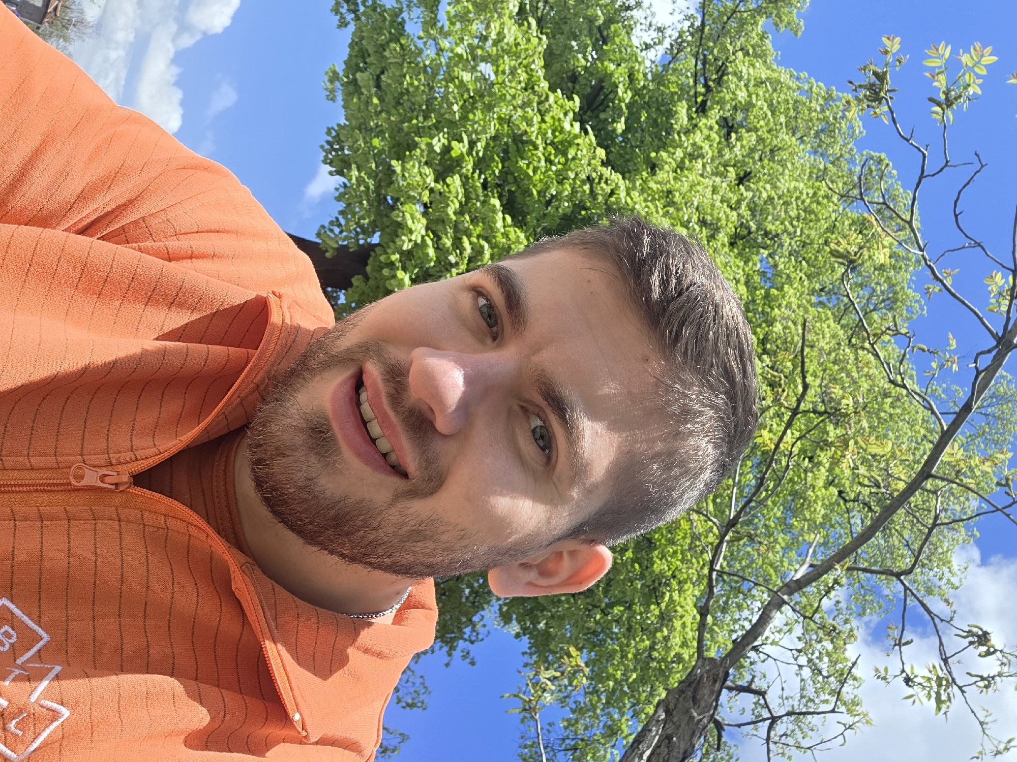 A man is posing for a selfie. He is wearing an orange zip-up hoodie. He has short brown hair and a beard. The background includes trees and a blue sky with some clouds. The location could be a park in Plovdiv, Bulgaria due to the presence of trees and a clear sky. The man is looking directly at the camera and smiling slightly. The image is taken from a low angle.
