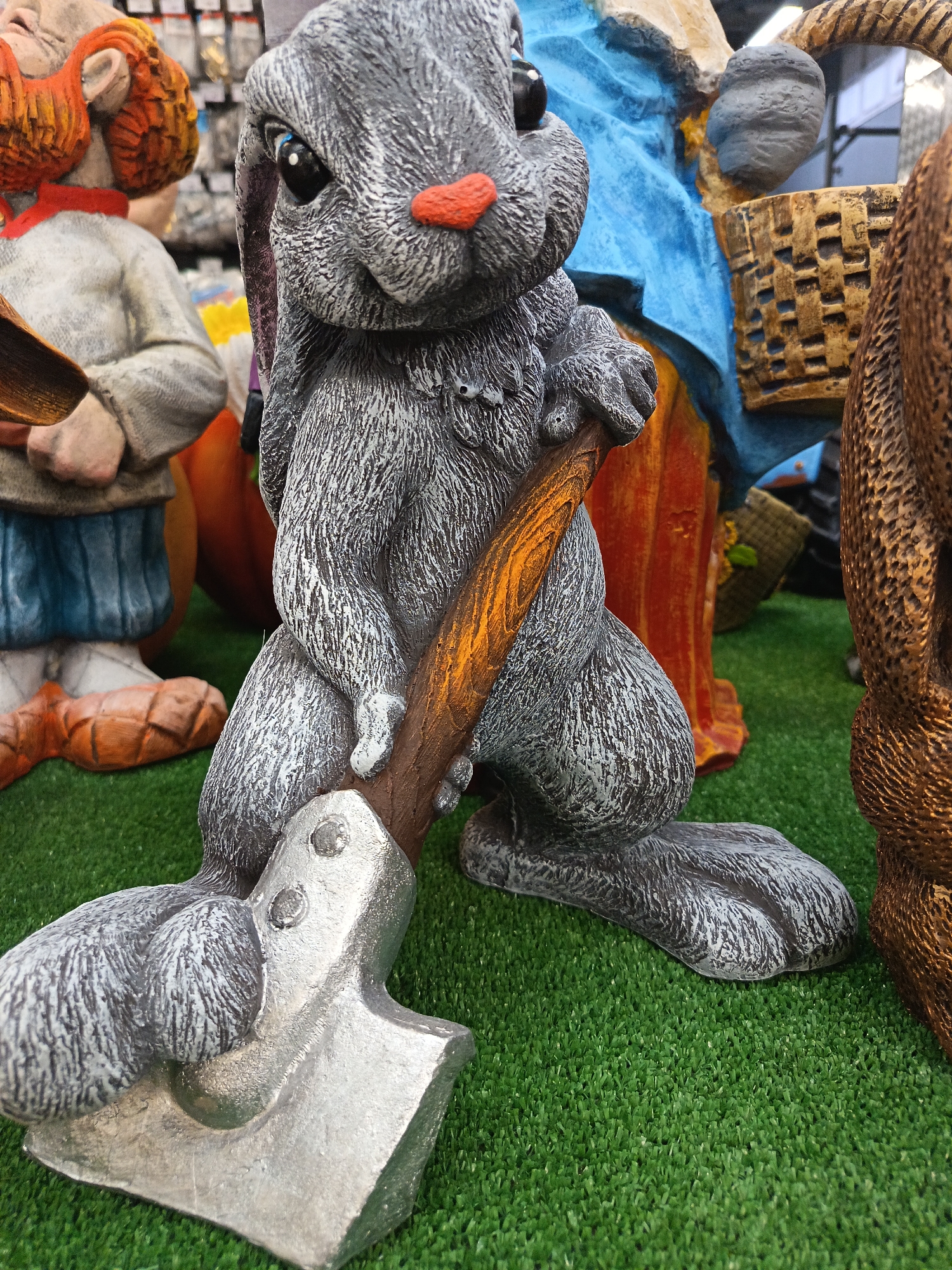 The image features a gray, textured garden statue of a rabbit holding an axe. The rabbit has a red nose and appears to be standing on a patch of artificial green grass. Other garden statues are visible in the background, including figures with orange hair and blue clothing, and a woven basket. The specific location in the city of Nizhnevartovsk, Russia, is not discernible from the image.