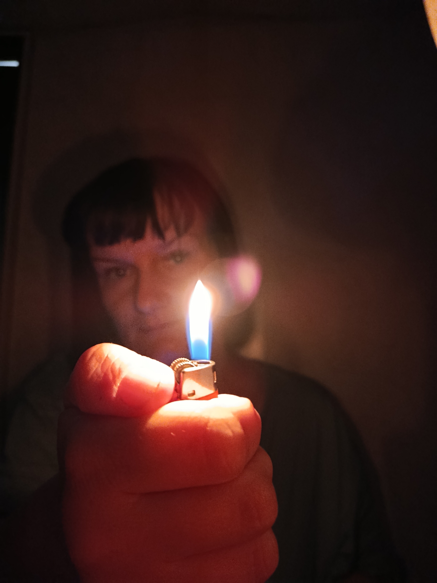 A person is holding a lit lighter in their right hand. The hand and lighter are close to the camera and well-lit by the flame. The person's face is blurred in the background, illuminated by the lighter. The background is dark and indistinct, appearing to be a wall. No other details suggest a specific location, and there is no evidence to indicate this photo was taken in Фоки, Russia.