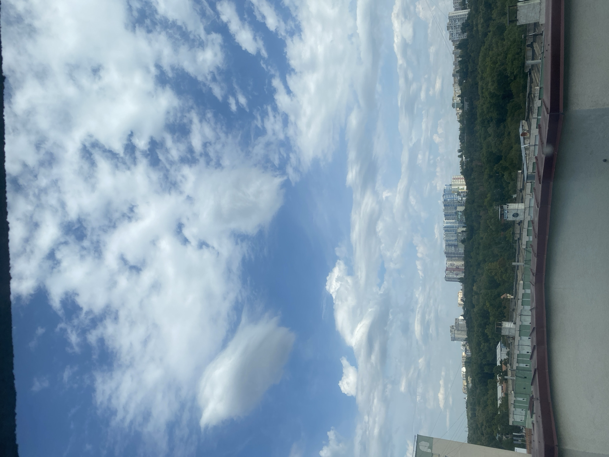 The image captures a panoramic view of a city skyline partially obscured by a foreground rooftop and a thick band of green trees. The sky is a vibrant blue, dotted with numerous white, fluffy clouds of varying sizes and shapes. In the distance, a cluster of modern, multi-story buildings stands out against the treeline, suggesting an urban or suburban setting. The buildings exhibit diverse architectural styles and colors, with some featuring light blue facades, others white, and some a mix of hues. The dense green foliage of trees covers a large portion of the middle ground, forming a natural buffer between the foreground and the distant cityscape. The foreground consists of a gray rooftop with a dark red trim. The overall impression is one of a bright, possibly sunny day, with the city nestled amidst a verdant landscape under a dynamic sky.