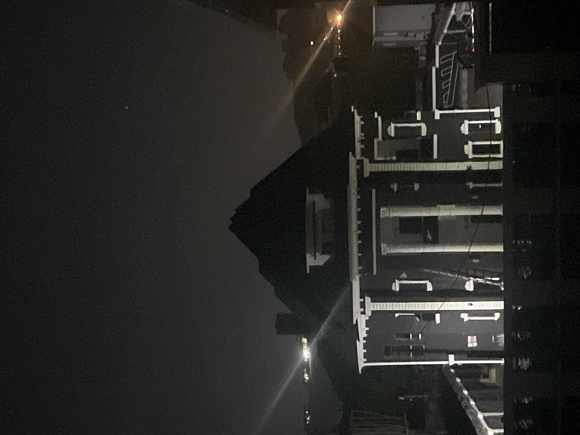 The image shows a nighttime view in Abuja, Nigeria. In the foreground, there is a building with multiple floors, illuminated with white lighting. The building appears to be adjacent to a body of water, possibly a river or lake, which is dark and mostly indistinguishable. Across the water, there is a pyramid-shaped landform, which might be a hill or mountain, silhouetted against the dark sky. There are a few faint lights scattered in the distance, suggesting the presence of other buildings or infrastructure. The overall impression is one of urban development near a natural landscape at night.
