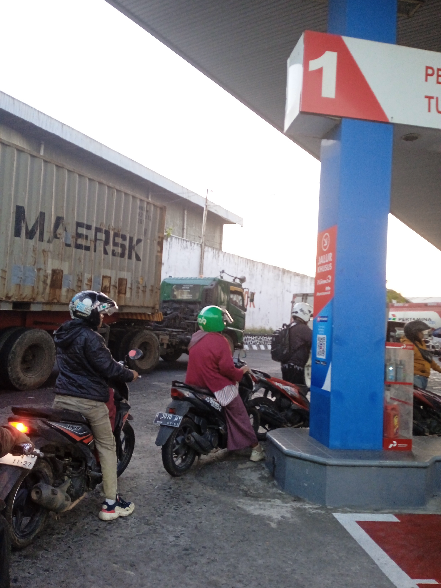 The image depicts a street scene in Semarang, Indonesia, likely at a gas station. In the foreground, two individuals are seated on motorcycles. The person on the left is wearing a black jacket, tan pants, and a white and black helmet. The person on the right wears a burgundy top and purple pants, also with a helmet. Behind them, a large, weathered MAERSK container truck dominates the middle ground, with a green truck visible to its right. A blue and red gas pump structure stands prominently in the right of the frame, displaying the number "1" on a red and white sign. A red sign with text and a QR code is attached to the blue structure. In the background, a white wall with some graffiti is visible. The ground is paved, and there are markings on the pavement, including a white arrow within a red section. The lighting suggests it is either early morning or late afternoon.