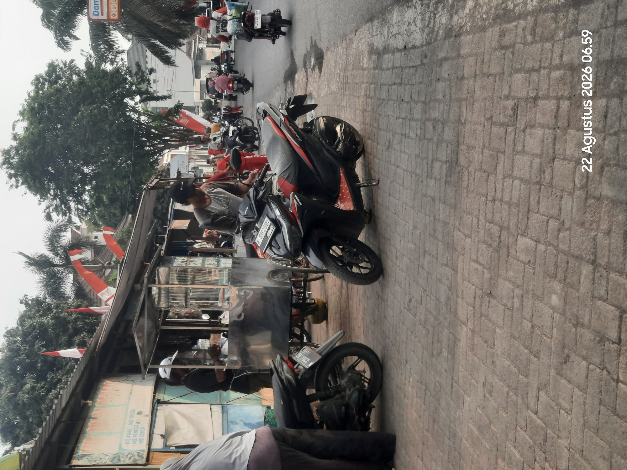 The image depicts a street scene in Pajarakan Kulon, Indonesia, on August 22, 2026, at 06:59. The foreground features a brick-paved sidewalk with two motorcycles parked on it. One motorcycle, a black Honda, is positioned in the center left, and another black motorcycle is partially visible on the right. A person, wearing dark clothing and a white cap, stands near a food cart on the left side of the image. The food cart appears to be made of metal and has a wheeled base. Behind the food cart, a building with a sloped roof is visible, adorned with Indonesian flags. Several other people are seen in the background, some on motorcycles, moving along the asphalt street. A tall green tree stands prominently in the middle ground. Various structures, including buildings and palm trees, line the street, suggesting an urban or semi-urban environment. The overall impression is of a bustling street scene, possibly in the morning.
