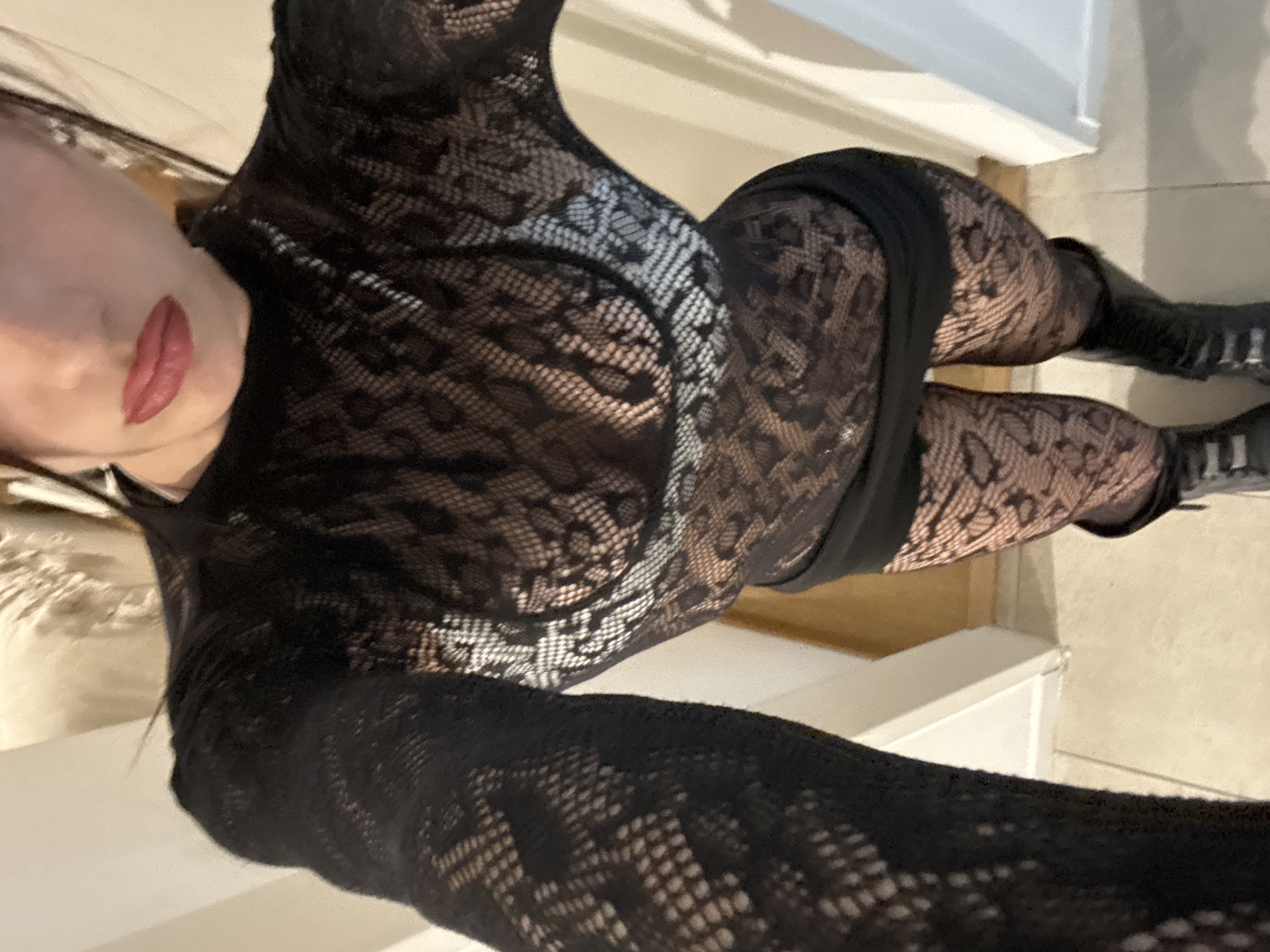 A woman wearing a black lace body suit, a black dress, black patterned tights, and black boots is posing indoors. The location is not identifiable as Buenos Aires, Argentina.