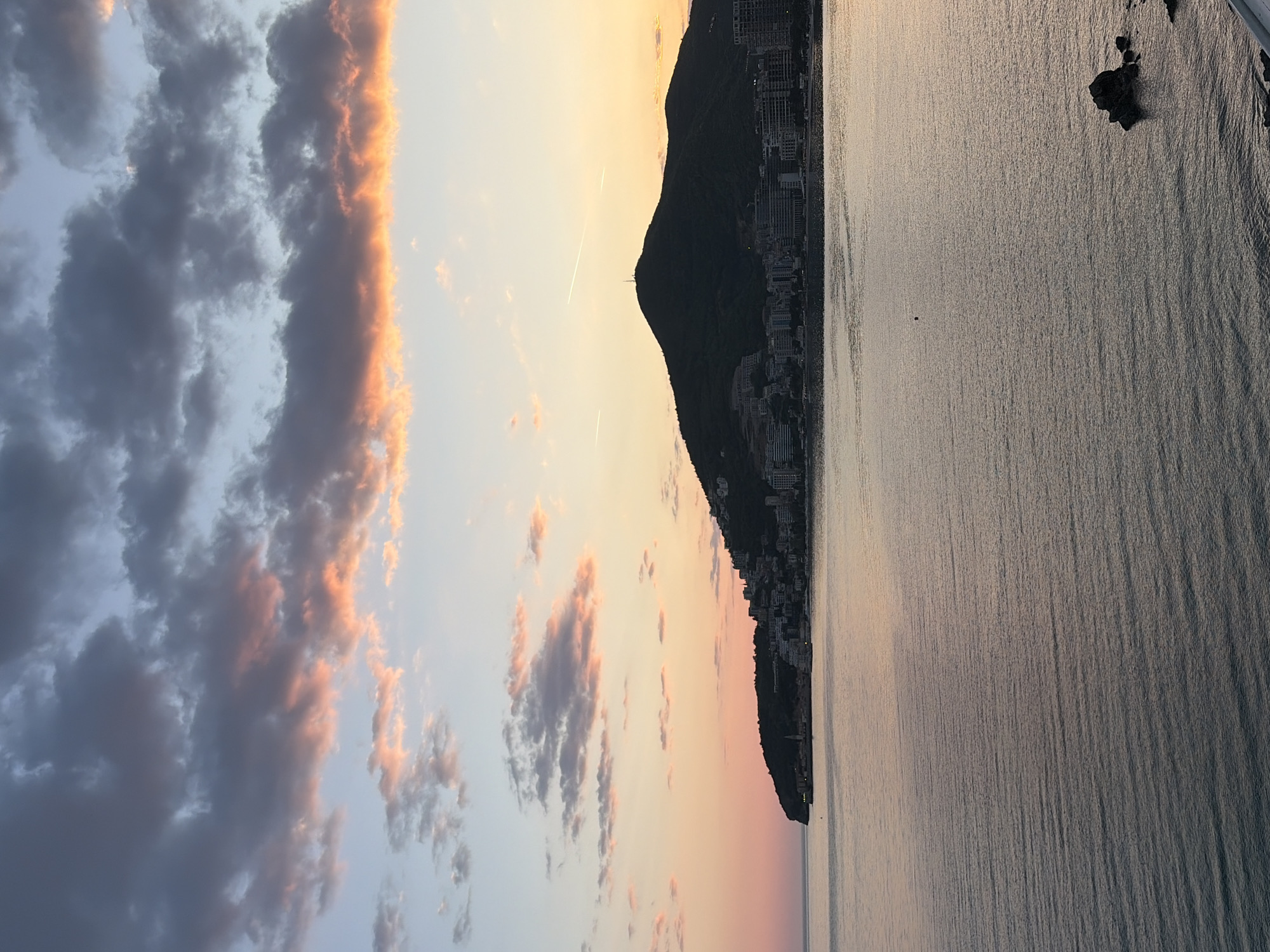 The image captures a scenic view in Budva, Montenegro, at what appears to be sunset or sunrise. The left side of the image shows a sky filled with clouds, some edged with a warm, pinkish-orange light. Two contrails are visible against the clear sky, which suggests some commercial aviation in the area. On the right, the still water reflects the colors of the sky, creating a mirror-like effect. A silhouette of a mountain or large hill runs vertically between the sky and the water. Buildings line the mountain's slope towards the water's edge, indicating urban development in the area. In the water, there are some visible rocks.