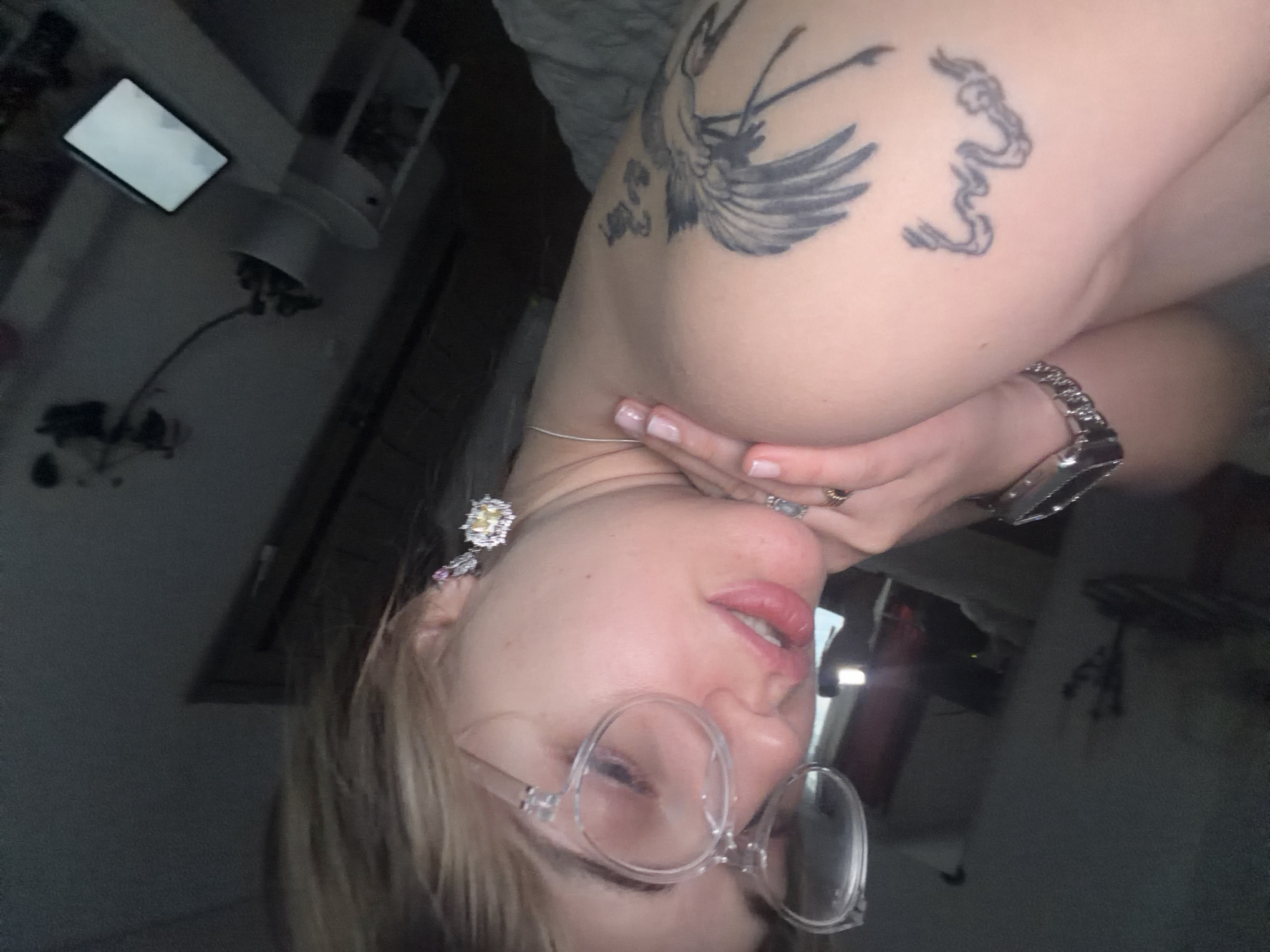 A selfie of a fair-skinned woman with blonde hair, wearing round glasses, an earring, a ring, and a watch. She has two tattoos on her right shoulder. The location is indoors. No specific information indicates the city of Большая Шильна, Russia.