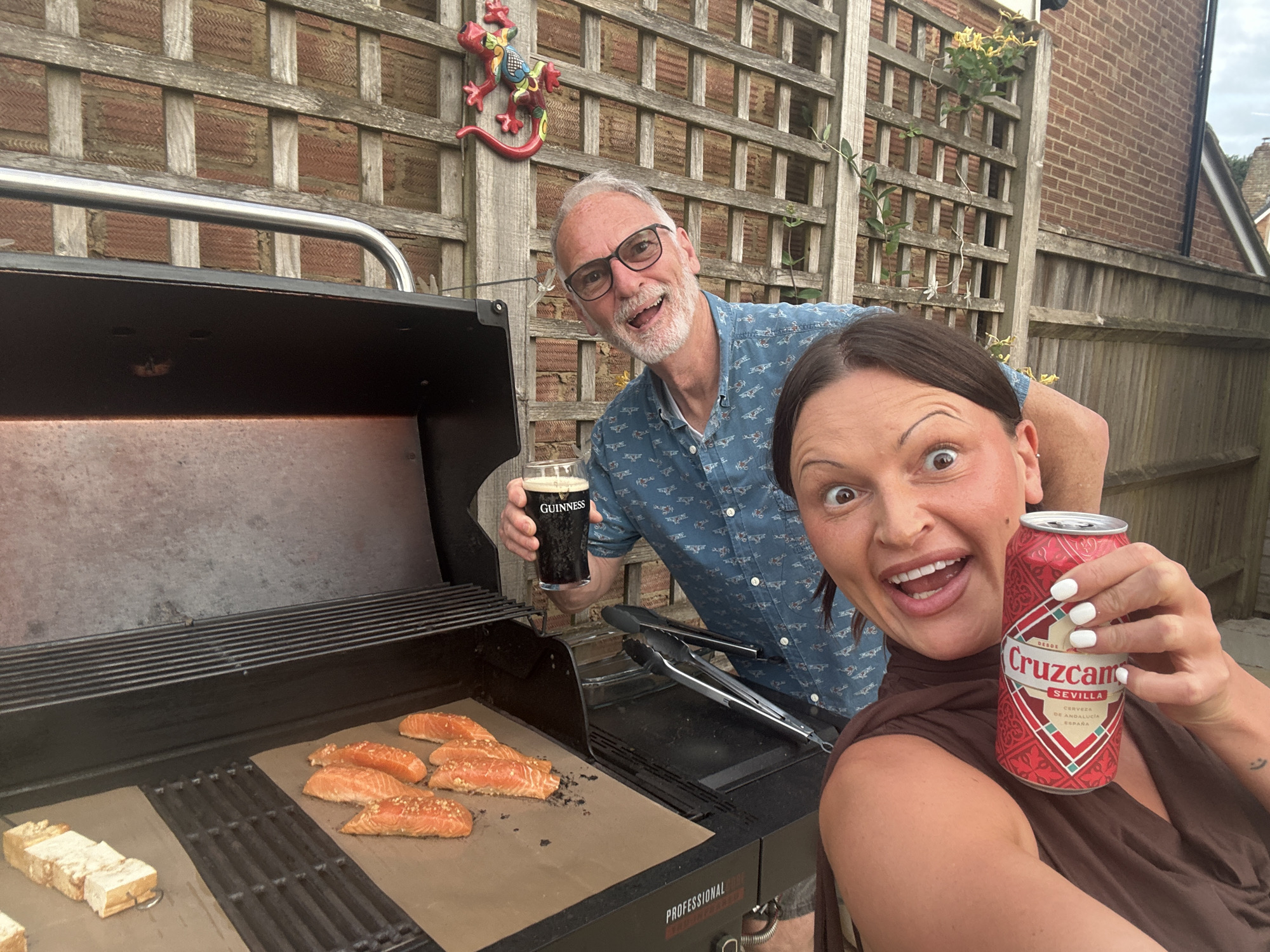 The image depicts two people in an outdoor setting, likely a backyard, with a grill in the foreground. A middle-aged man with glasses is holding a pint of Guinness beer and smiling. Beside him, a woman is holding a can of Cruzcampo Sevilla beer and smiling widely at the camera. The grill has pieces of salmon cooking on it. The background shows a fence, brickwork, and some greenery. There are no explicit indicators or clues to suggest the city of Tunbridge Wells is the setting of this photo.