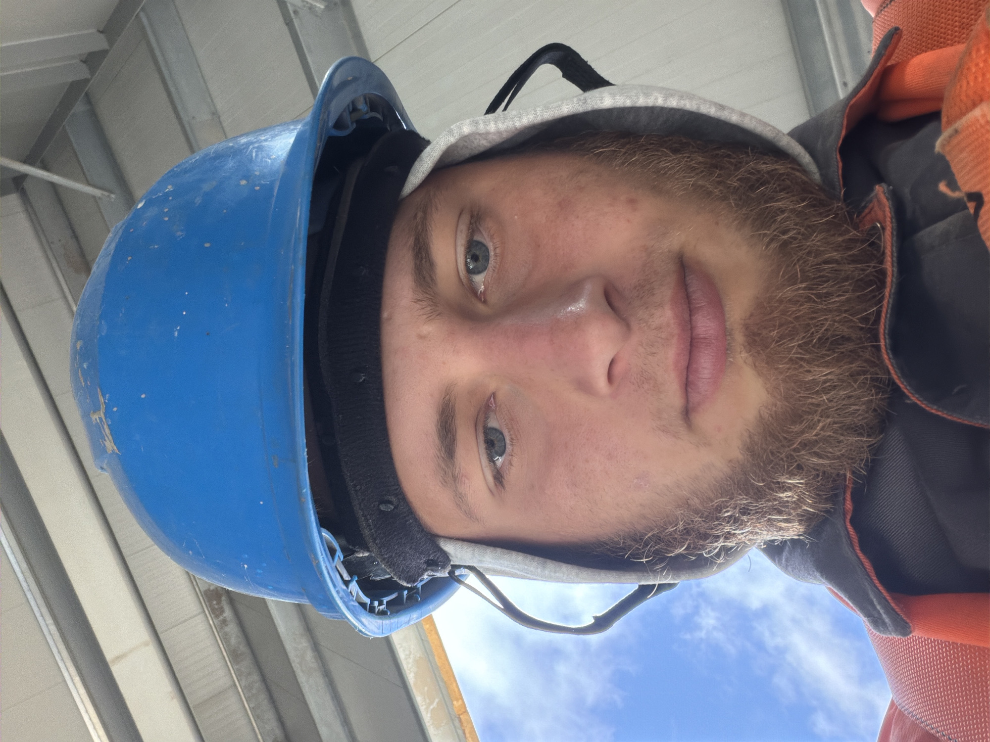A low-angle, close-up, selfie-style photograph shows a young Caucasian man wearing a blue hard hat and an orange safety vest. He has light skin, blue eyes, and a full, reddish-brown beard. His hair is not visible under the hard hat. He is wearing a grey hooded sweatshirt under the vest, with the hood pulled up and framing his face. The background appears to be an interior with overhead beams, and a sliver of blue sky with white clouds is visible to the left. The image is taken from a perspective looking up at the man. The location is identified as Stróża, Poland.