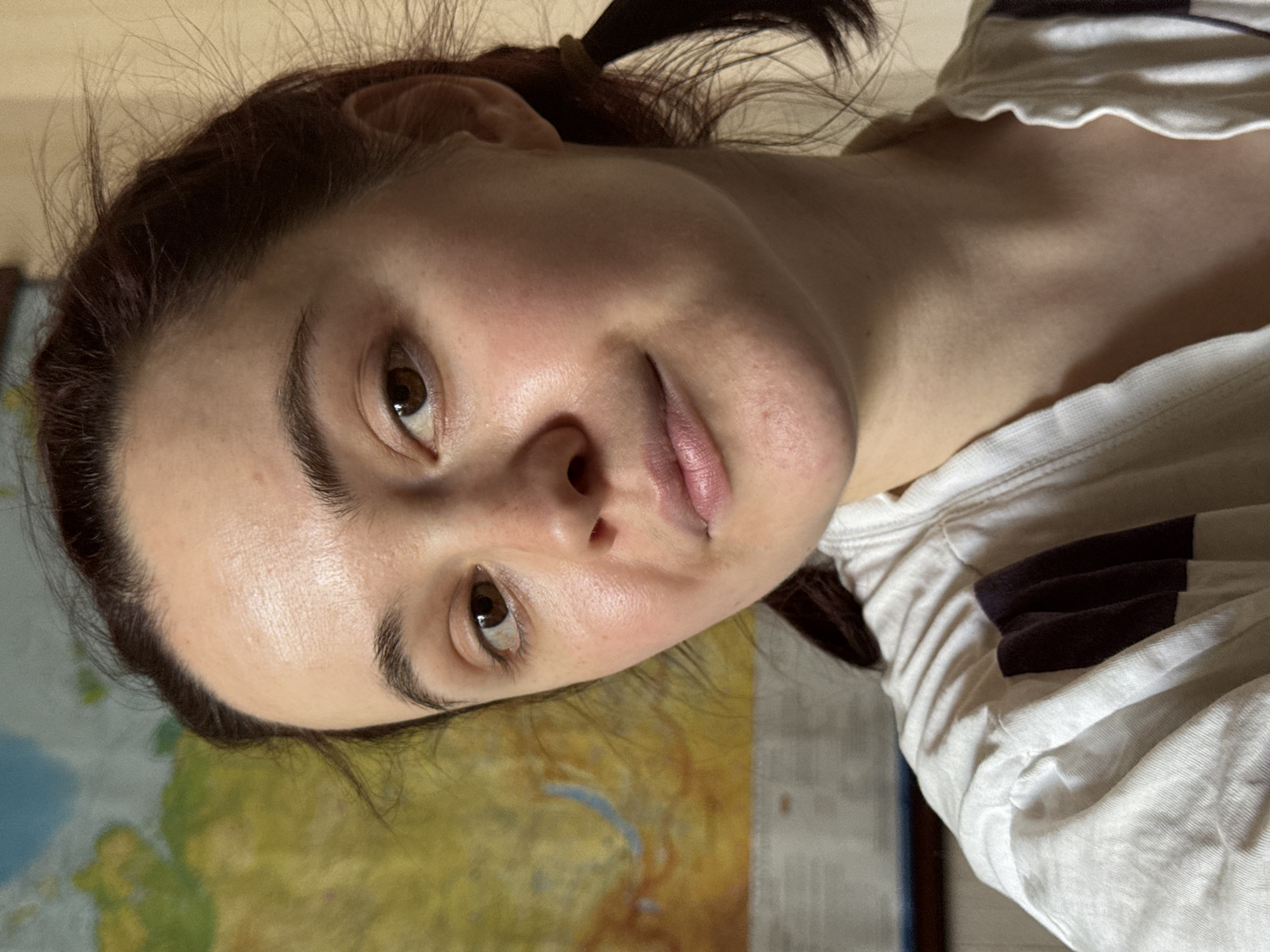 A close-up, slightly rotated portrait shot of a woman with fair skin, brown hair pulled back into a small ponytail, and brown eyes. She is looking directly at the camera with a slight smile. She is wearing a white t-shirt with dark stripes. A portion of a map with yellow and blue coloration is visible in the background. The image location cannot be determined by the provided information.