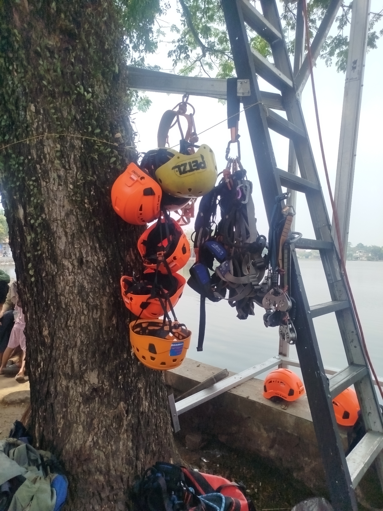 This image depicts an outdoor scene in Depok, Indonesia, near a body of water, likely a lake or river, given the presence of a distant shoreline with vegetation and buildings. In the foreground, a large tree trunk is covered in moss and vines. Hanging from the tree are several orange and yellow climbing helmets, each with straps. Adjacent to the tree is a tall metal ladder, leaning against what appears to be a wooden structure or platform. Several harnesses and carabiners are suspended from the ladder. Two orange helmets are placed on the platform below the ladder. In the background, a hazy view of the water and distant landscape is visible. Two individuals are partially visible in the lower left quadrant of the image, one wearing a pink skirt and the other a green garment. Several bags and backpacks are scattered on the ground near the base of the tree. The overall impression is of an outdoor adventure or recreational activity setup.