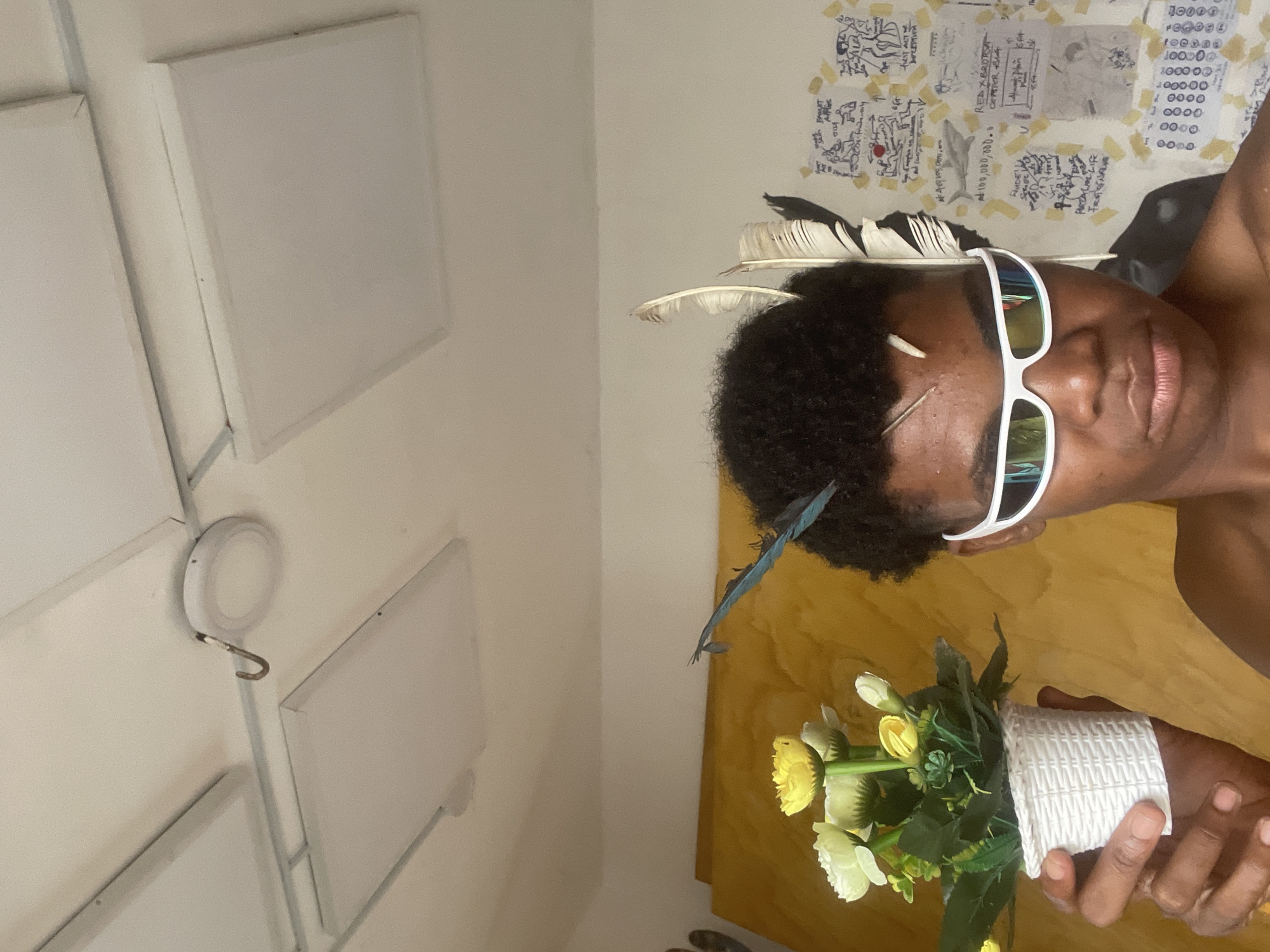 A person with dark curly hair, wearing white sunglasses and several feathers in their hair, holds a small white pot with artificial yellow and white flowers. The person is shirtless and looking directly at the camera with a neutral expression. Behind them, a wall is covered with various drawings, notes, and yellow tape. The ceiling has recessed lighting panels and a hook. The scene appears to be indoors, possibly in a room in Ado, Nigeria.
