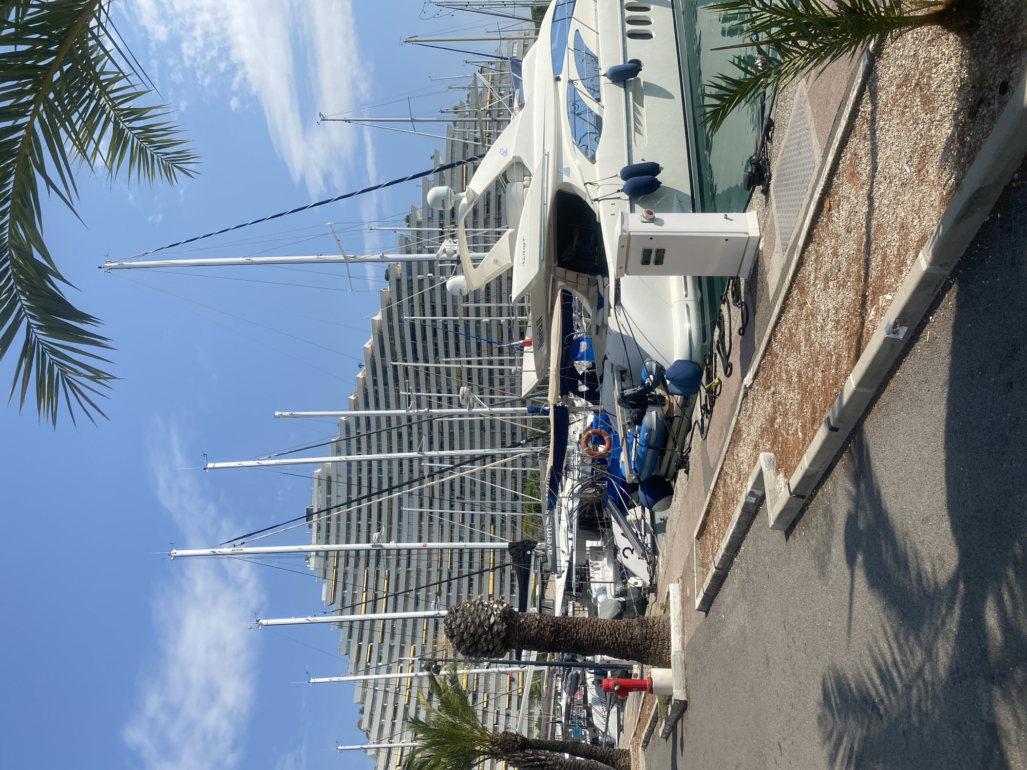 The image depicts a marina in Villeneuve-Loubet, France. In the foreground, a paved walkway runs alongside the water, casting shadows from palm fronds. A red fire hydrant is visible to the left, next to a palm tree. The marina itself is filled with numerous sailboats and a large white yacht. In the background, a distinctive, stepped apartment building with many balconies and yellow awnings dominates the skyline. The sky is blue with scattered white clouds. The scene appears to be a sunny day.