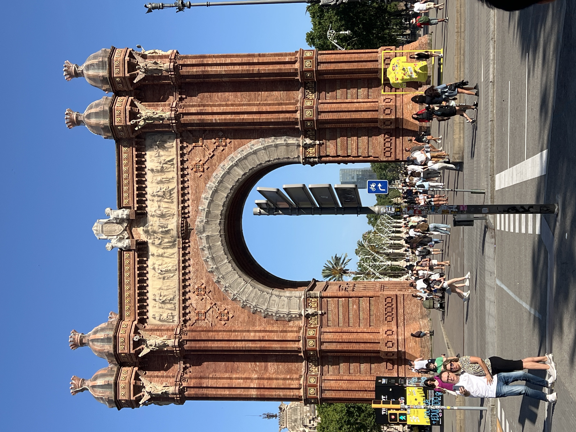 The image captures the Arc de Triomf in Barcelona, Spain, under a clear, bright blue sky. The prominent central subject is the red brick Arc de Triomf, a monumental structure featuring a large central archway. Its facade is adorned with stone relief carvings, including friezes depicting multiple figures above the arch and winged figures positioned on the upper corners of the structure. The archway's upper sections are also embellished with decorative patterns and a crest on the far right. The arch is flanked by four octagonal towers, each topped with a distinct cupola and a crown-like finial.
The foreground consists of an asphalt road with a white pedestrian crossing. To the left, a traffic light displays a red signal, and a blue sign with a white bicycle icon is visible. A couple is centered in the immediate foreground, posing for a selfie; the man wears a white t-shirt and blue jeans, while the woman wears a leopard-print top and a black skirt. Numerous other pedestrians are present on the wide paved areas surrounding the arch and along the tree-lined avenue extending through it.
On the right side of the frame, adjacent to the base of the arch, a large yellow display shaped like a t-shirt or jersey is erected on a metal frame. Palm trees and other green foliage are visible to the sides of and through the arch. In the distant background, through the archway, modern high-rise buildings are discernible, indicating an urban environment. The scene is illuminated by strong sunlight, casting distinct shadows and suggesting daytime.