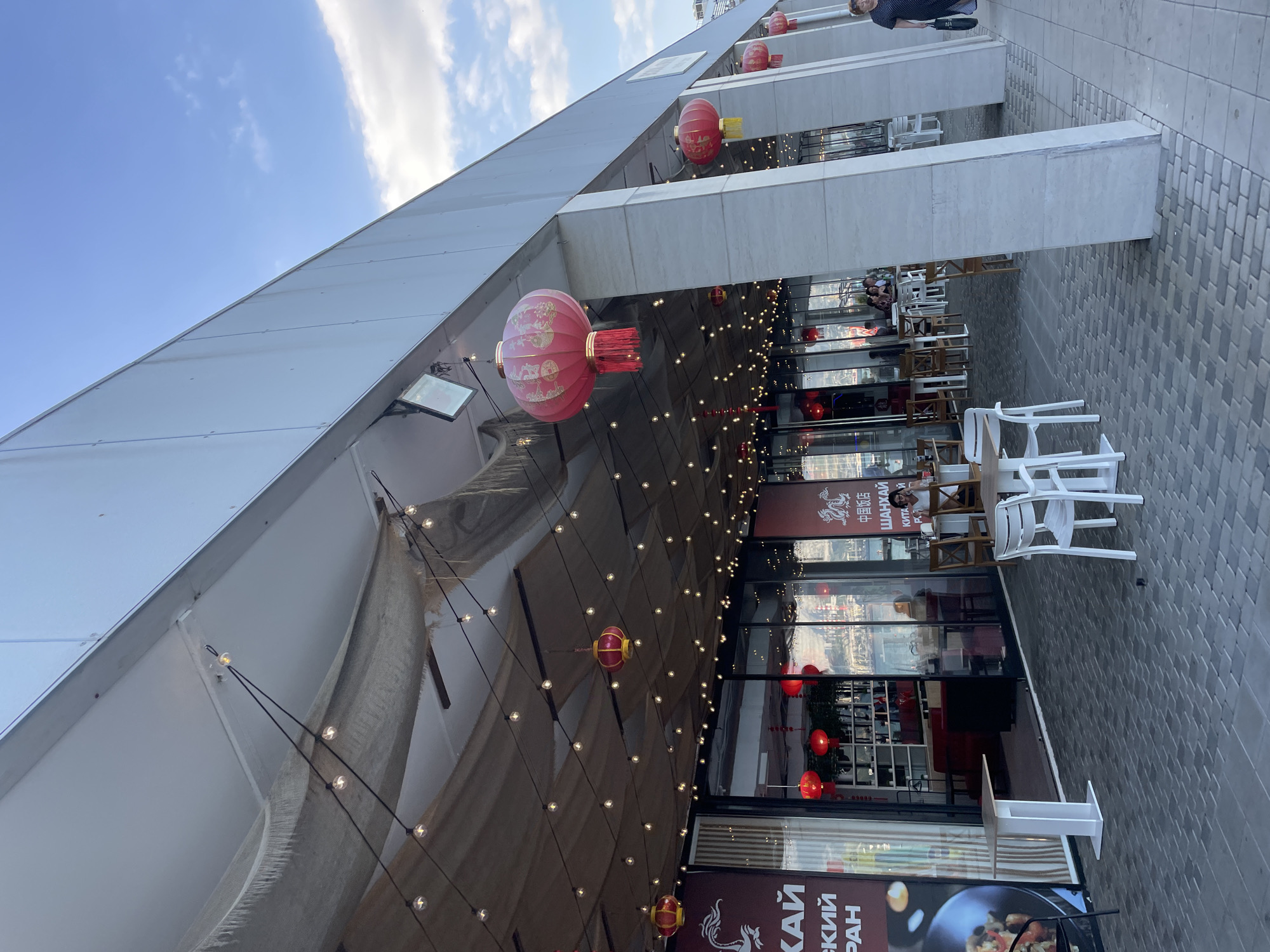 The image captures an outdoor seating area of a Chinese restaurant, possibly during daytime. The setting appears to be urban, with a paved walkway leading to the establishment. Overhead, a white roof structure extends outwards, adorned with strings of small, warm-toned lights and several large, red Chinese lanterns. The lanterns are decorated with intricate patterns.
The restaurant's interior is visible through large glass windows, revealing tables and chairs, as well as shelving units. Some of these tables are set up outside, with white plastic chairs and wooden chairs arranged around them. People are seated at some of the outdoor tables, engaged in conversation and dining.
The sky above is partly cloudy, suggesting a pleasant, possibly warm day. The architecture of the building is modern, with clean lines and a white facade. There is visible text on the building's signage. One sign clearly displays Chinese characters ("中国饭店" which translates to "Chinese Restaurant") alongside Russian text ("ШАНХАЙ" which translates to "Shanghai"). Another sign features the Russian phrase "КАЙСКИЙ ПРАН", also related to the restaurant's theme. The overall atmosphere seems relaxed and inviting, suggesting a leisurely dining experience.