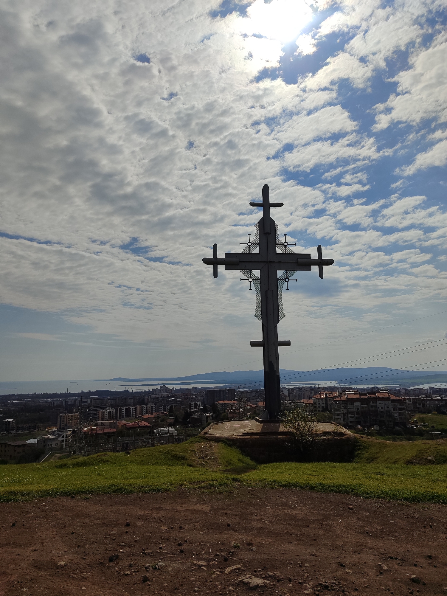 The image shows a large metal cross atop a hill in Burgas, Bulgaria. The cross is mounted on a concrete base, surrounded by green grass. The foreground is a dirt ground. Beyond the cross, a cityscape stretches towards the sea, with buildings of various sizes and colors visible. The sky is partly cloudy with the sun visible. In the distance, there are mountains across the sea. Power lines are also visible in the background.