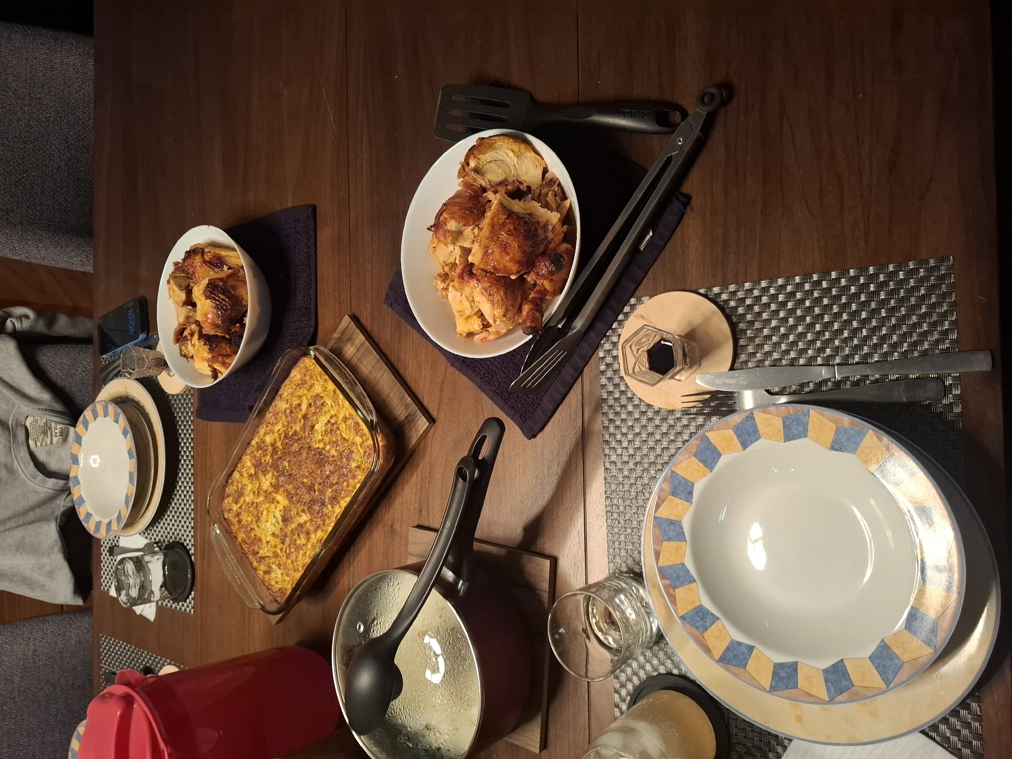 The image depicts a dinner table set for a meal. In the foreground, a place setting includes a decorative plate and silverware. To the left, a pot with a lid and ladle is visible, suggesting a warm dish. A glass of amber liquid with ice is also present. In the background, a glass baking dish contains a golden-brown casserole. A white bowl filled with carved roasted chicken sits on a dark blue cloth. A red pitcher is positioned to the left of the casserole. The table is made of dark wood, and a woven placemat is under the plate and silverware. The lighting suggests an indoor setting, possibly during the evening. The image does not provide any specific visual cues to determine the location within Huixquilucan de Degollado, Mexico.