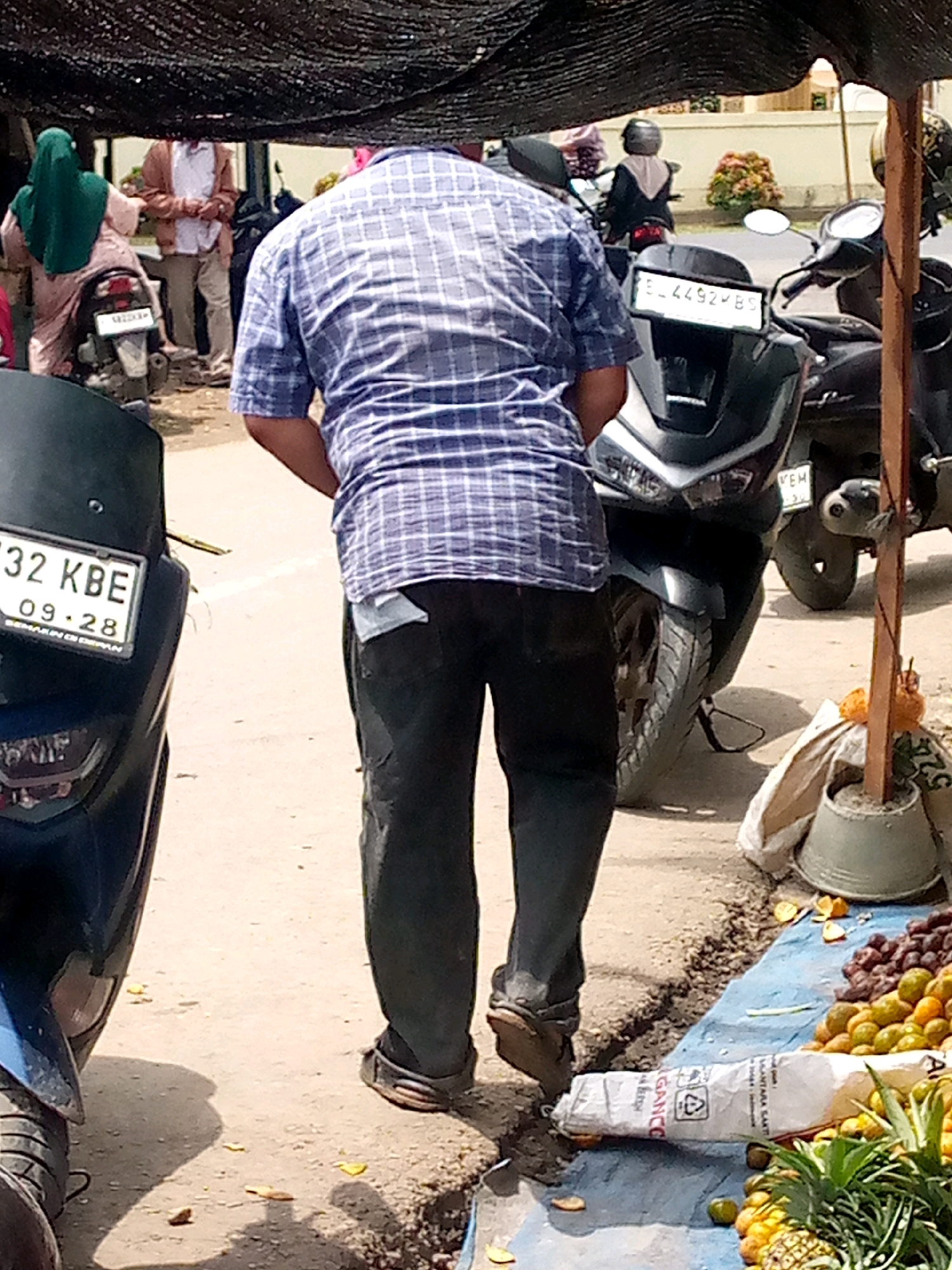 In Keudeteupinpunti, Indonesia, a man wearing a blue plaid shirt and dark pants stands in a street market setting. He is facing away from the camera, with his right hand in his pants pocket. To his left, a dark blue motorcycle with a license plate reading "32 KBE 09-28" is parked. To his right, a motorcycle with a license plate reading "EL 4492 YBS" is visible. In the foreground, a blue tarp displays a variety of fruits, including oranges, grapes, and pineapples. A white bag lies on the tarp next to the fruits. The ground is paved with asphalt and dirt. A wooden post with some orange decorations stands to the right of the tarp. Other people and motorcycles are visible in the background, indicating a busy market environment.