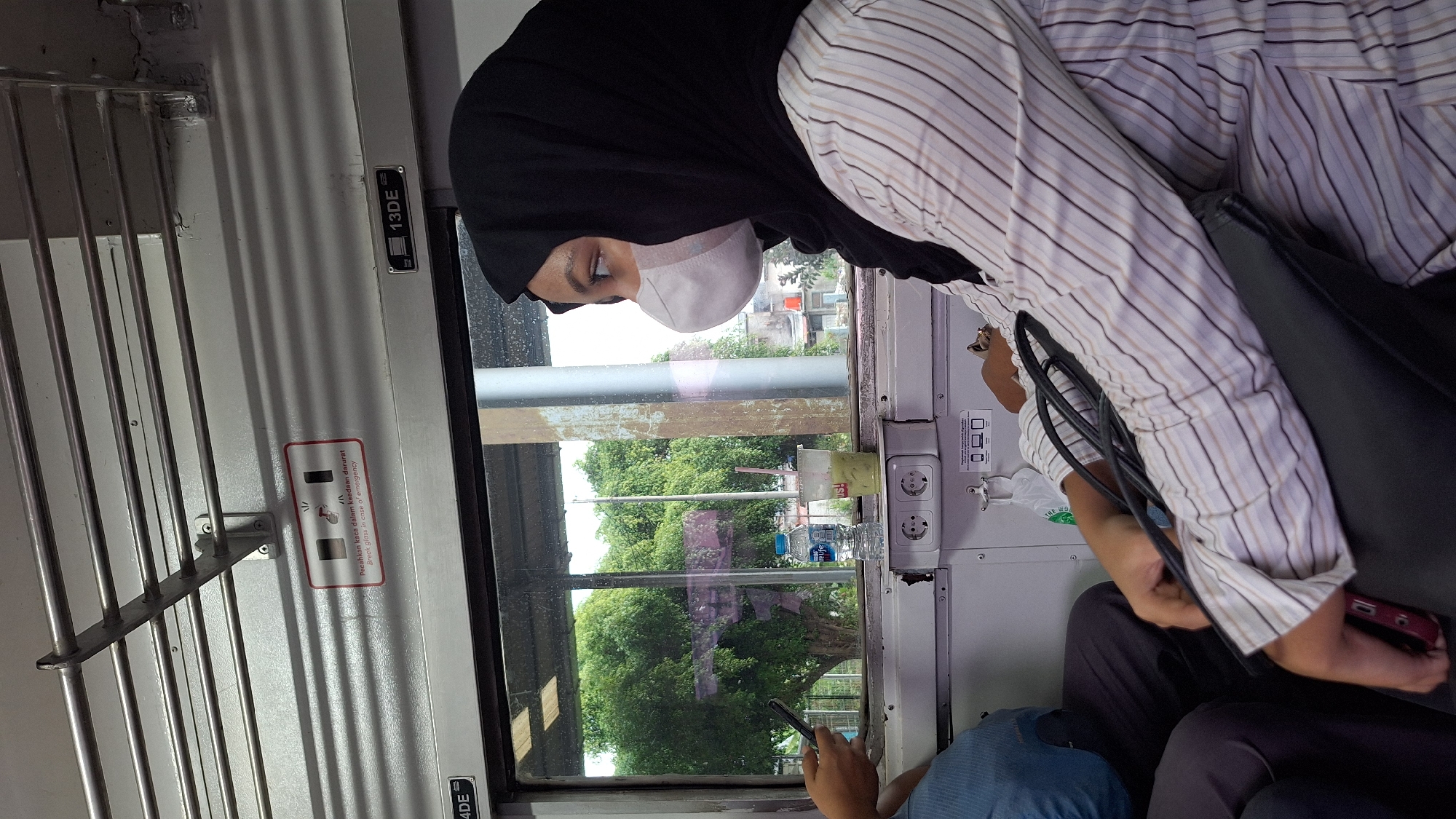 The image captures an interior view of a train carriage, likely in the city of Karawang, Indonesia, based on the contextual information. In the foreground, a person wearing a black hijab and a white and brown striped shirt is seated, looking out the window. They are also wearing a white face mask. Their arms are resting on a black bag. To the left, another person is partially visible, wearing a blue shirt and dark pants, holding a smartphone. Through the window, lush green trees and some buildings are visible, suggesting a semi-urban or suburban landscape. On the train wall, there are emergency instructions with text in Indonesian and English. Electrical outlets are also visible on the wall below the window.