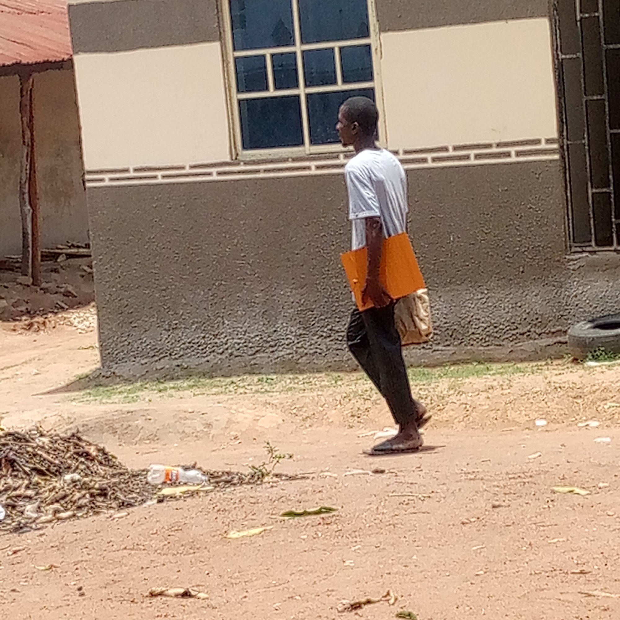 A man is walking on a dirt road in an unidentified urban location. He is carrying an orange folder under his arm and a tan bag over his shoulder. He is wearing a white shirt and dark pants. The background includes a building with a window and a pile of debris next to a plastic bottle. A tire also rests against the building wall.