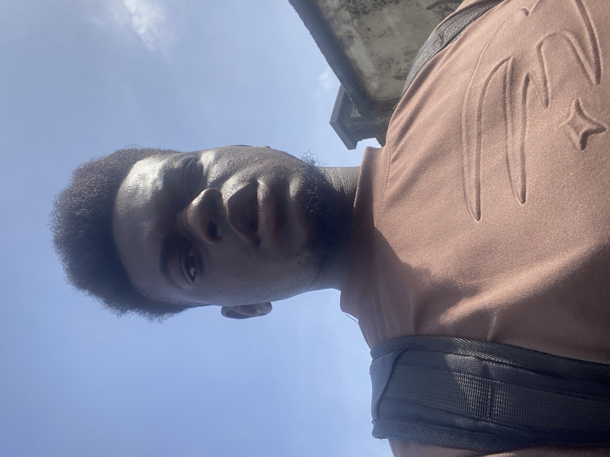 A low-angle, upward-facing shot of a Black man outdoors. He is wearing a brown t-shirt with raised design elements and a black strap, likely from a bag, is visible across his shoulder. The sky is light blue and slightly cloudy. A concrete structure is partially visible in the background. The location is not identifiable.