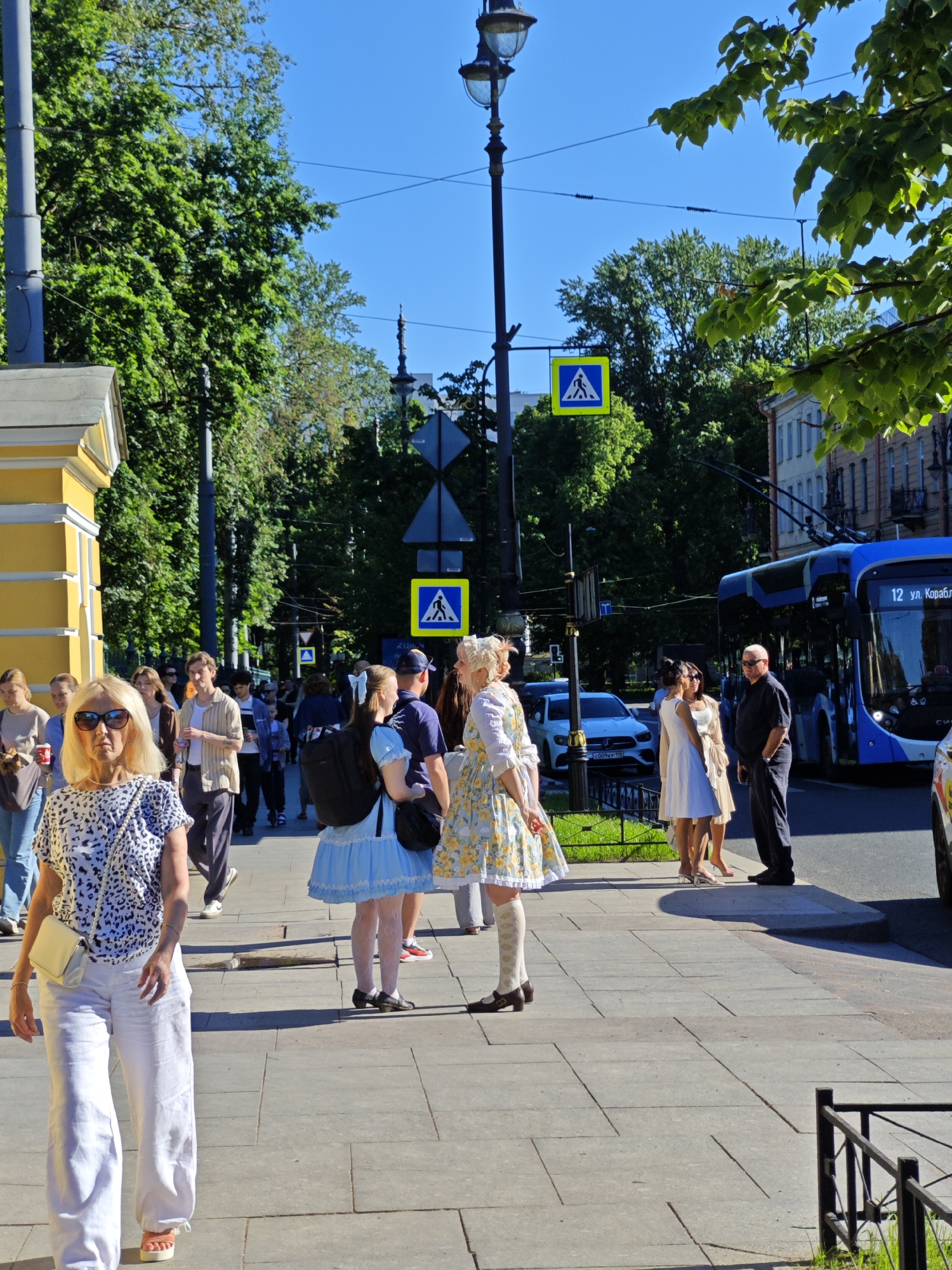 This is a detailed image of an outdoor urban scene on a clear, sunny day in Saint Petersburg, Russia. The perspective is from a paved sidewalk looking down a tree-lined street.
In the foreground, a blonde woman with sunglasses, wearing a black and white leopard print short-sleeved top and white wide-leg pants, walks towards the viewer on the left side of the sidewalk. She carries a light yellow shoulder bag. To her right, further back, are several pedestrians. Notably, two individuals in distinct outfits are visible: one woman in a light blue dress with a large white bow in her hair, white lace stockings, and black shoes, carrying a black backpack; and another woman in a yellow and white floral patterned dress with an ornate white headpiece, white socks, and brown shoes. They stand near a man in a dark blue polo shirt and cap.
The street features dark grey light poles with ornate lanterns, from which multiple overhead power lines extend. Two yellow square pedestrian crossing signs with blue backgrounds and white pedestrian symbols are prominently displayed, one nearer on the right and another further down the street. The left side of the scene is dominated by a bright yellow building with white trim. In the mid-ground, a dark blue modern bus with "12 ул. Кораблестроителей" (12 ul. Korablestroiteley) displayed on its front approaches on the road. Near the bus, a man in a black shirt and dark pants stands conversing with two women in light-colored dresses (one white, one beige). A white Mercedes-Benz car is partially visible behind them.
The street is lined with numerous tall, lush green trees, creating a dense canopy of foliage on both sides. In the background, classical-style multi-story buildings with various window treatments are visible. A black metal fence separates the sidewalk from a small strip of grass in some areas. Long shadows are cast across the sidewalk, indicating bright sunlight. The sky is a clear, deep blue.