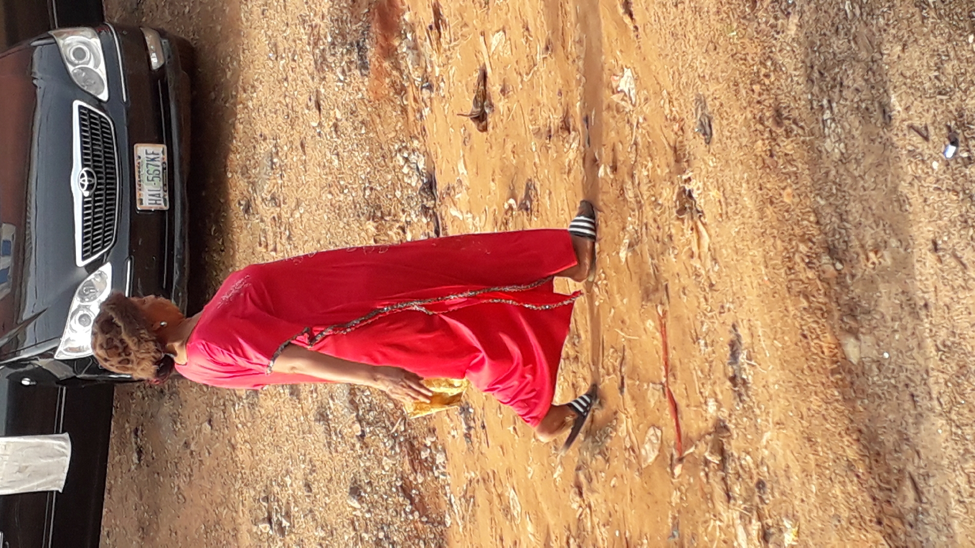 A woman in a red dress and striped sandals stands on a dirt road next to a black car. The license plate of the car reads "HAL-557KF." The location appears to be Nkpor, Nigeria.
