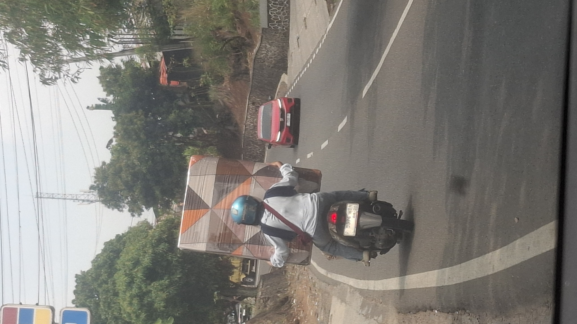 The image shows a person riding a motorcycle on a road in Sirnagalih, Indonesia. The rider is wearing a blue helmet and is carrying a large, rectangular object wrapped in patterned plastic on their lap. The object appears to be a mattress or similar item. The motorcycle has a license plate that reads "F 5374 HDN". A red car is visible further down the road, and trees and a stone wall line the side of the road. In the background, a radio tower and power lines are visible against a hazy sky. The road has white lane markings.