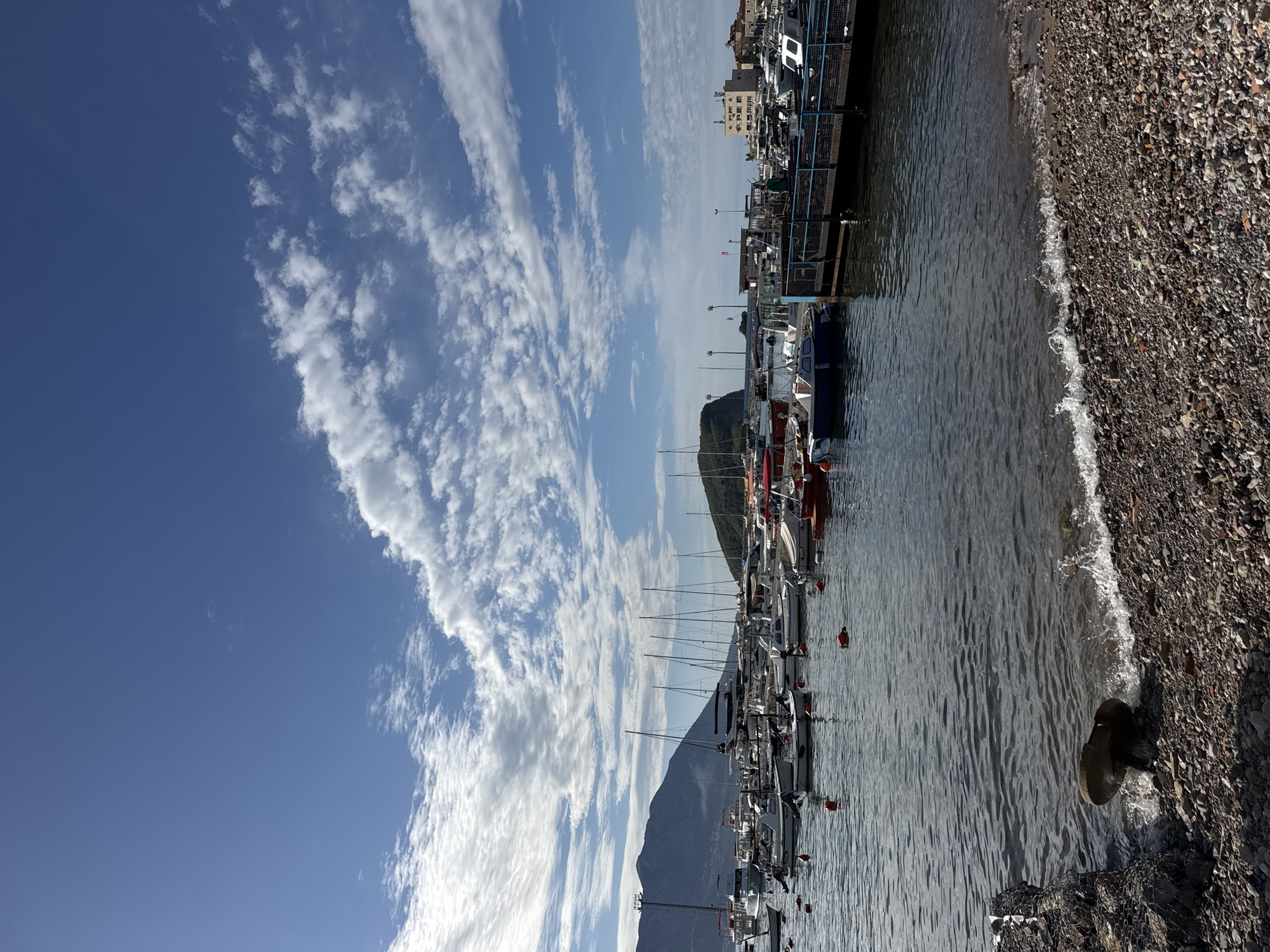 The image depicts a harbor scene in Budva, Montenegro, with a clear blue sky streaked with wispy clouds. A mountain looms in the background, partially obscured by the masts of numerous sailboats moored in the harbor. The water reflects the sky, with gentle waves lapping against a pebbled beach. A concrete pier runs along the side of the water with buildings on the pier, indicating the edge of the city. Some dark shapes appear at the edge of the pebbles and in the water that look to be rocks.