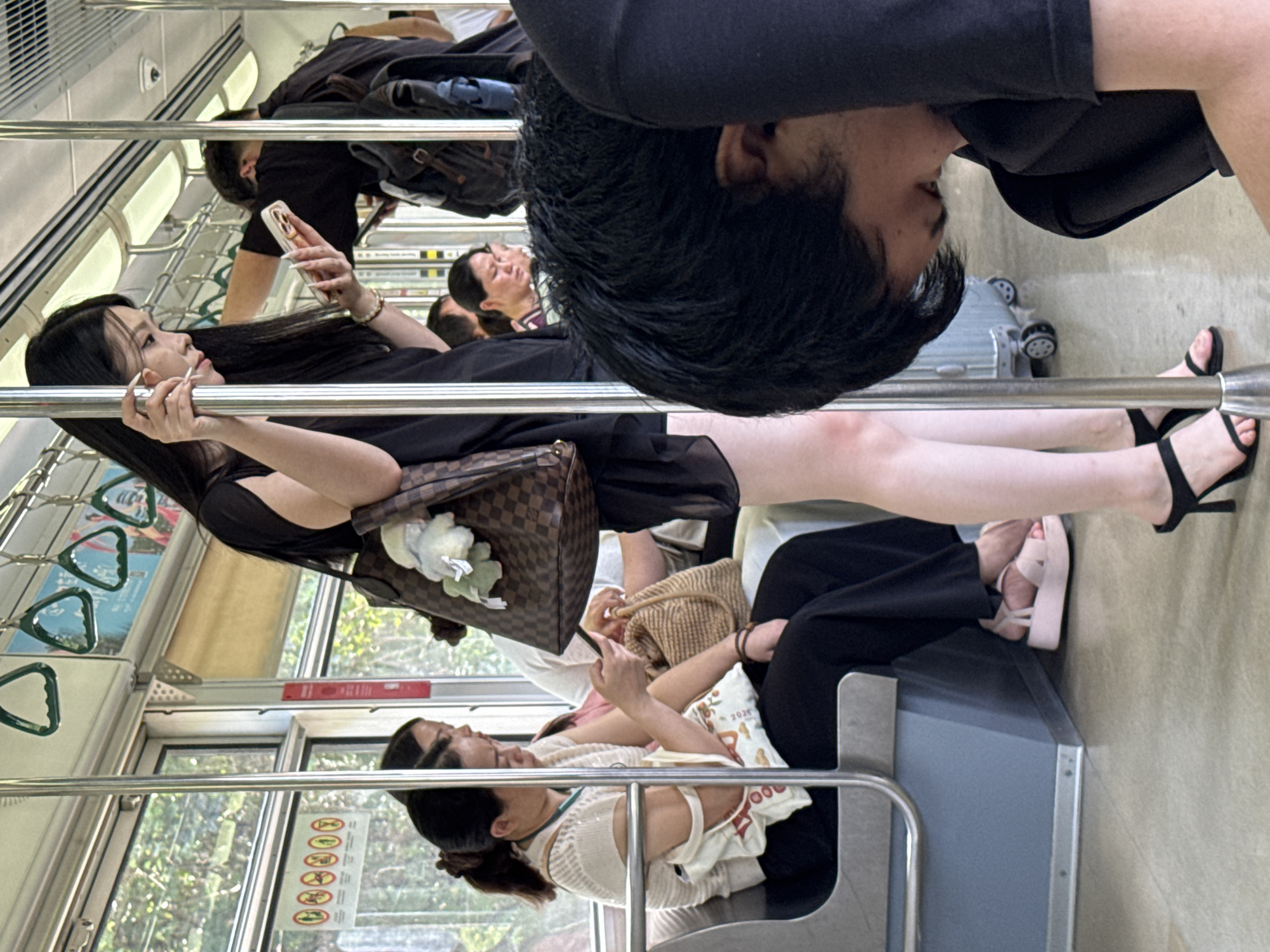 The image shows the interior of a crowded subway train in Yuzhong District, China. In the foreground, a person with dark, tousled hair is hunched over, obscuring their face. To the left, a young woman with long black hair, wearing a black dress and black high heels, stands holding onto a pole and looking at her phone. She carries a large designer handbag with a floral embellishment. Further to the left, two women are seated on the bench. One woman, wearing a cream-colored top and black pants, is looking at her phone. Next to her, another woman is partially visible. The subway car has metal poles for passengers to hold onto, overhead hand straps, and windows revealing a glimpse of green trees outside. The floor is a light-colored, textured material. The overall atmosphere suggests a busy commute.