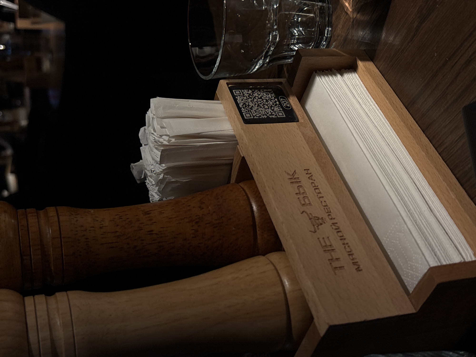 The image shows a tabletop setting in what appears to be a restaurant. A wooden napkin holder with a QR code sticker and the text "THE БыК МЯСНОЙ РЕСТОРАН" (The Bull Steak Restaurant) is visible. The napkin holder contains both folded paper napkins and stacked white napkins. A clear glass water glass sits behind the napkins. Below the napkins are wooden salt and pepper shakers. The background is dark and blurry, suggesting an interior setting. The restaurant is likely located in Moscow, Russia, based on the Russian text on the napkin holder.