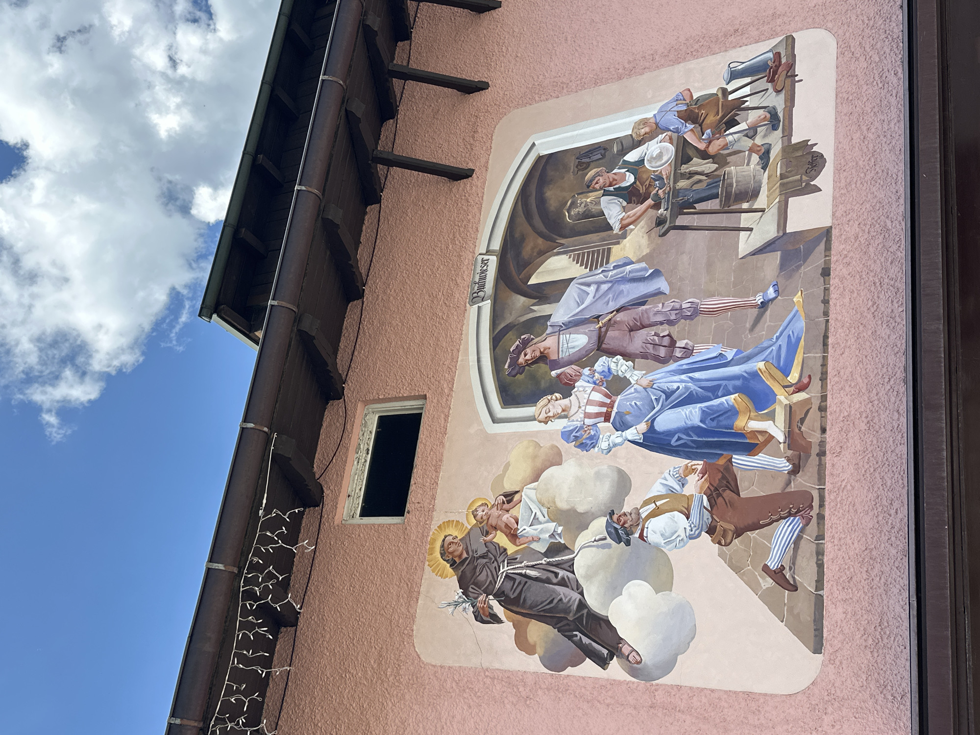 This is a detailed mural painted on a pink stucco building facade in Garmisch-Partenkirchen, Germany. The mural depicts a historical scene with several figures. On the left, a robed figure, likely a religious saint, is depicted with a halo and holding a lily, with a baby on his arm. He is surrounded by clouds, and below him, a man kneels in prayer or supplication, dressed in traditional attire with striped leggings and a leather pouch.
To the right of the saint, a man and a woman stand side-by-side. The woman wears an elaborate blue dress with puffed sleeves and a red and white striped bodice. The man is dressed in a long, flowing purple cloak over a tunic and brown breeches.
Further to the right, an arched entryway leads to an interior scene with two men working at a workbench. One man, wearing an apron, is crafting an object, possibly pottery or metalwork, on a stand. A younger boy, also in an apron, sits nearby, observing or assisting. Tools, a bucket, and a pitcher are visible on and around the workbench. The words "Buchwieser" are written in an arch above this scene, and "Treffer" is inscribed on a scroll-like element below the workbench. The building's brown gutters and wooden roof supports are visible above the mural, against a blue sky with scattered white clouds. The overall scene evokes a sense of history and craftsmanship.