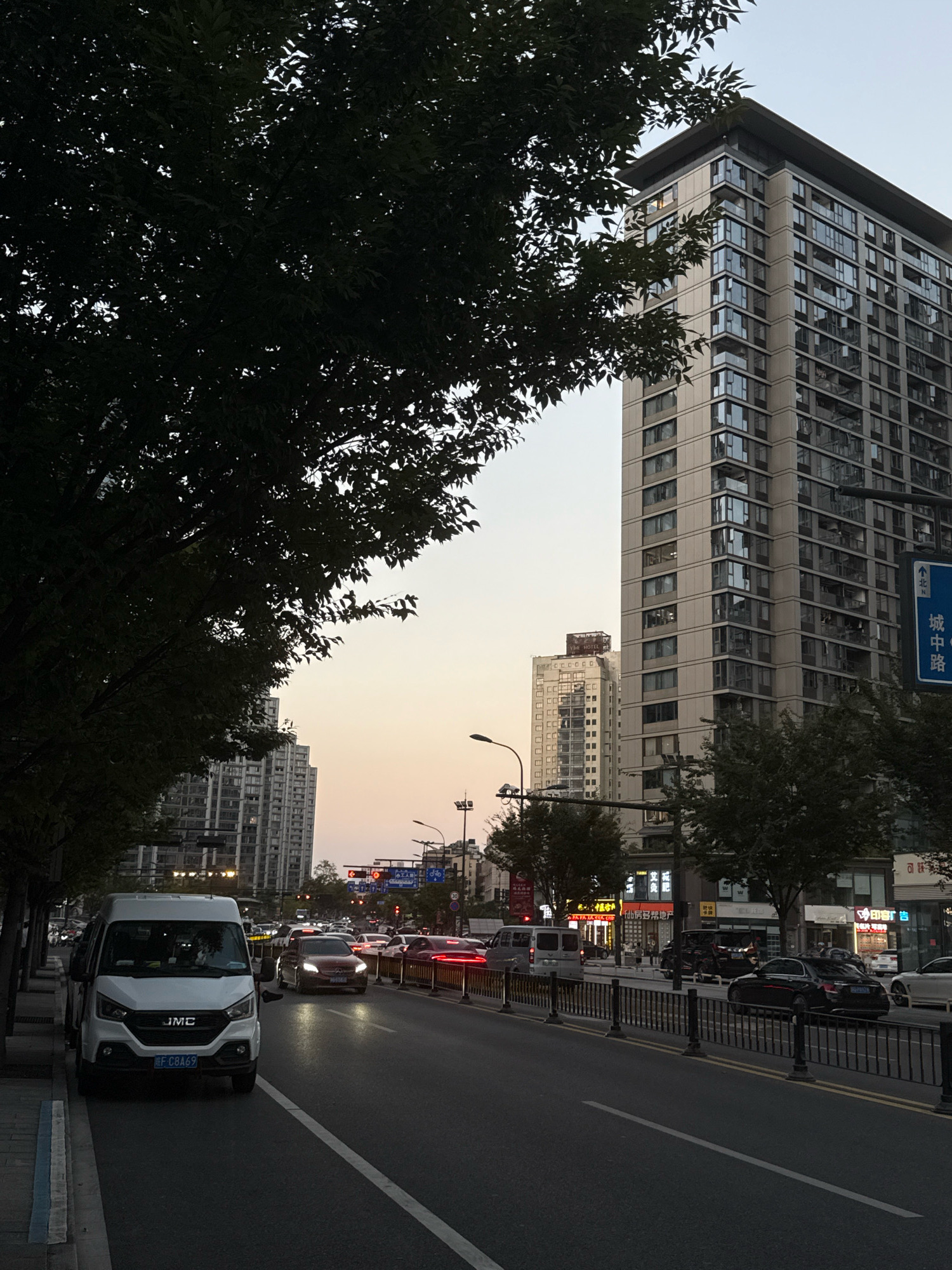The image depicts a street scene in Yiwu, China, characterized by a wide asphalt road with multiple lanes of traffic. Tall residential buildings with numerous windows dominate the background on the right side of the street. In the foreground, a white JMC van is parked on the left side of the road, facing away from the viewer. Several other vehicles, including cars and vans, are traveling on the road. The sky exhibits a warm, twilight hue, suggesting it is either early morning or late evening. Trees with dark foliage line the left side of the street, partially obscuring the view of the buildings. Traffic lights and street lamps are visible, indicating an urban environment. The presence of shops and businesses with illuminated signs along the street further reinforces the urban setting.