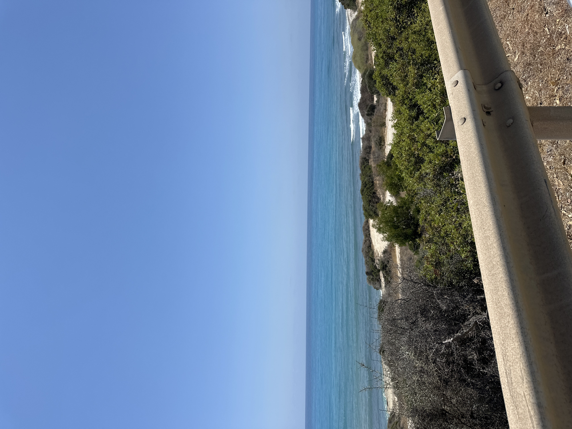 The image shows a coastal scene under a clear blue sky. In the foreground, a dusty, beige metal guardrail runs horizontally across the bottom of the frame. Beyond the guardrail, a dry, brown-toned landscape with sparse vegetation, including dry bushes and some green shrubs, leads to a sandy area. Further in the background, the turquoise ocean stretches to the horizon, with gentle waves breaking on the shore. The sea meets the sky in a sharp, clear line. The location is identified as the city of Aphrodite Hills, Cyprus, which suggests a Mediterranean coastal environment. There are no people or animals visible in the image. The overall impression is one of a bright, sunny day at the seaside.