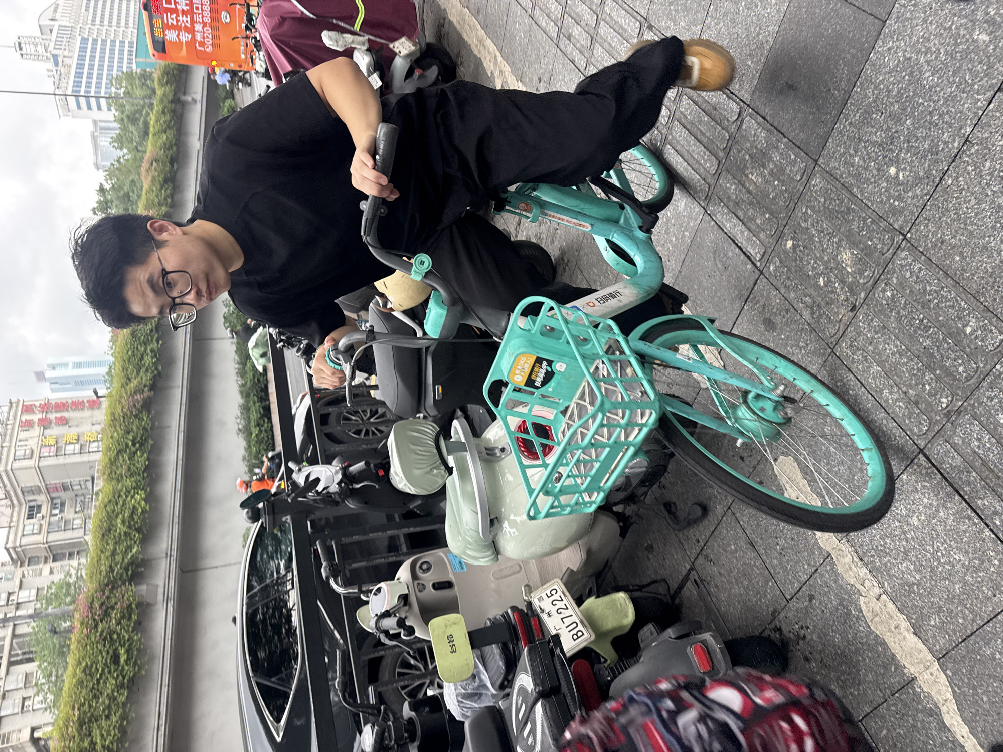 The image shows a man in a black shirt and black pants, wearing glasses, riding a light blue bicycle. The bicycle has a basket in the front. He is on a paved area with tactile paving indicators. In the background, there is a black car and several parked mopeds and scooters. Buildings are visible in the background, suggesting an urban environment. Text in Chinese indicates the location is in Tianhe District, Guangzhou, China. A sign is partially visible on an orange bus with Chinese characters and a phone number: 9020-8888.