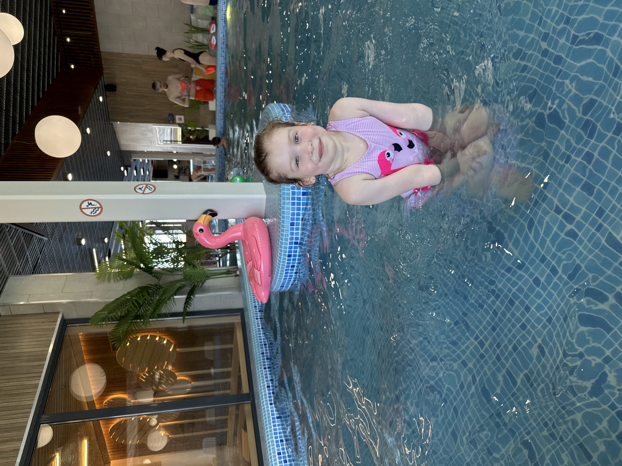 A young girl in a pink flamingo swimsuit is sitting in a pool. The pool has light blue tiles and water. There is a pink flamingo float next to her. Behind her, there are other people in the pool and lounge chairs in the background. There is also a potted palm tree. The location is most likely an indoor swimming pool. The location cannot be verified as Votkinsk, Russia, based on the visual information provided.