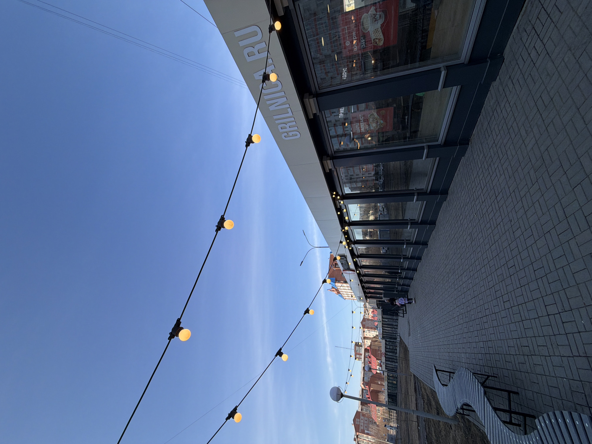 The image depicts an outdoor scene in Krasnoyarsk, Russia. A modern building with a storefront bearing the sign "GRILLNICA RU" runs along a paved walkway. Strung with lights, the building's design features large windows. The walkway includes benches. In the distance, residential buildings with red roofs are visible. The sky above is clear and blue, with electrical wires stretching across it. In the distance, two individuals appear to be seated on a bench.