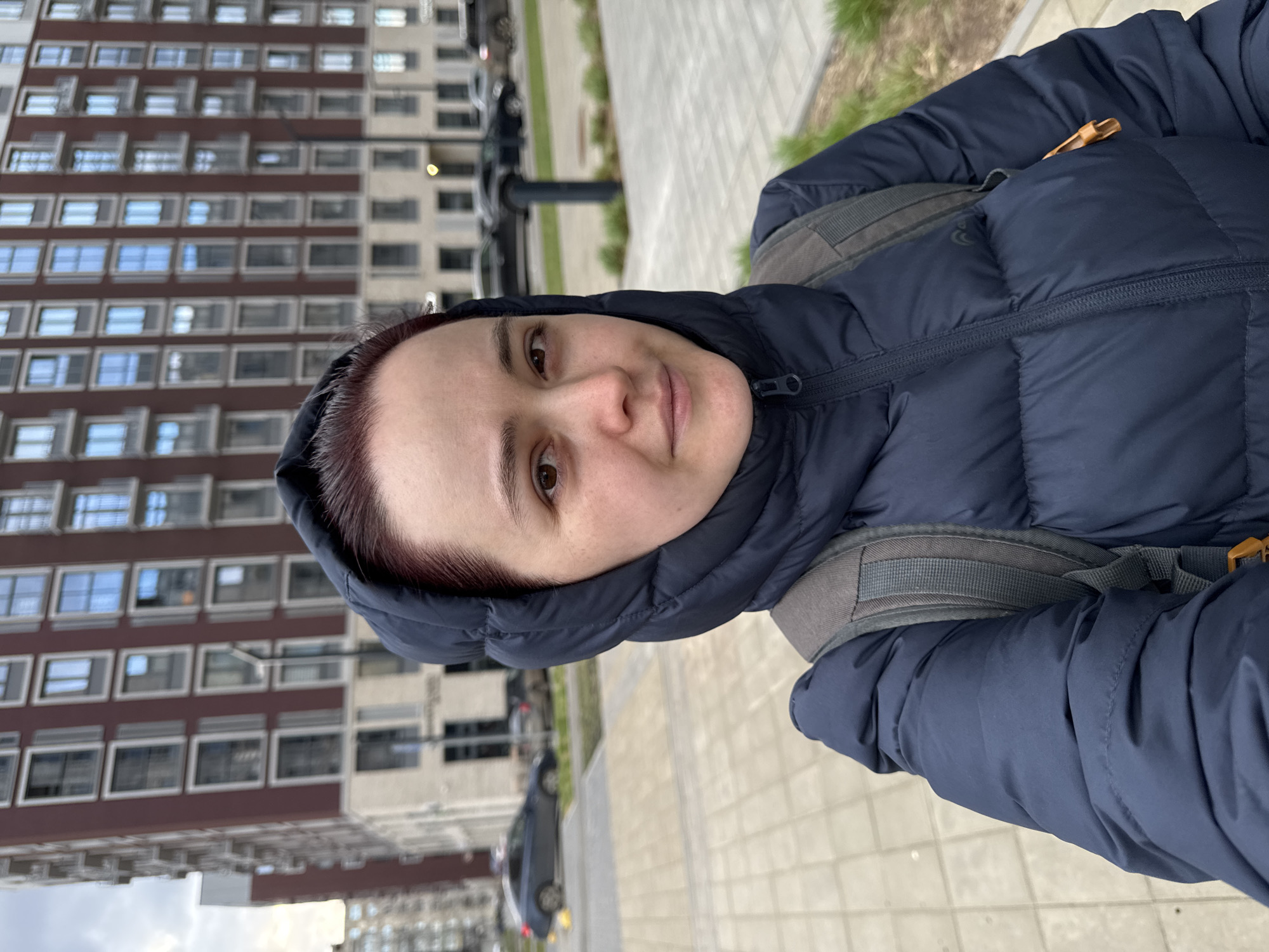 The image shows a woman taking a selfie outdoors in Izhevsk, Russia. She is positioned in the foreground, facing the camera with a neutral to slight smile. She has dark reddish-brown hair, dark eyes, and is wearing a dark blue hooded puffer jacket with the hood pulled up. Grey straps of a backpack are visible on her shoulders beneath the jacket.
The background reveals an urban or residential setting. Behind her are modern multi-story buildings, one prominently featuring a brown facade with numerous rectangular windows with white frames, and another with a lighter beige/white color. A paved sidewalk extends to the right, and what appears to be a street with parked cars is visible further in the background. Sparse green patches of grass can be seen alongside the pavement. The lighting suggests it is daytime, possibly overcast, with soft, diffused light and no harsh shadows. There is no visible text in the image.