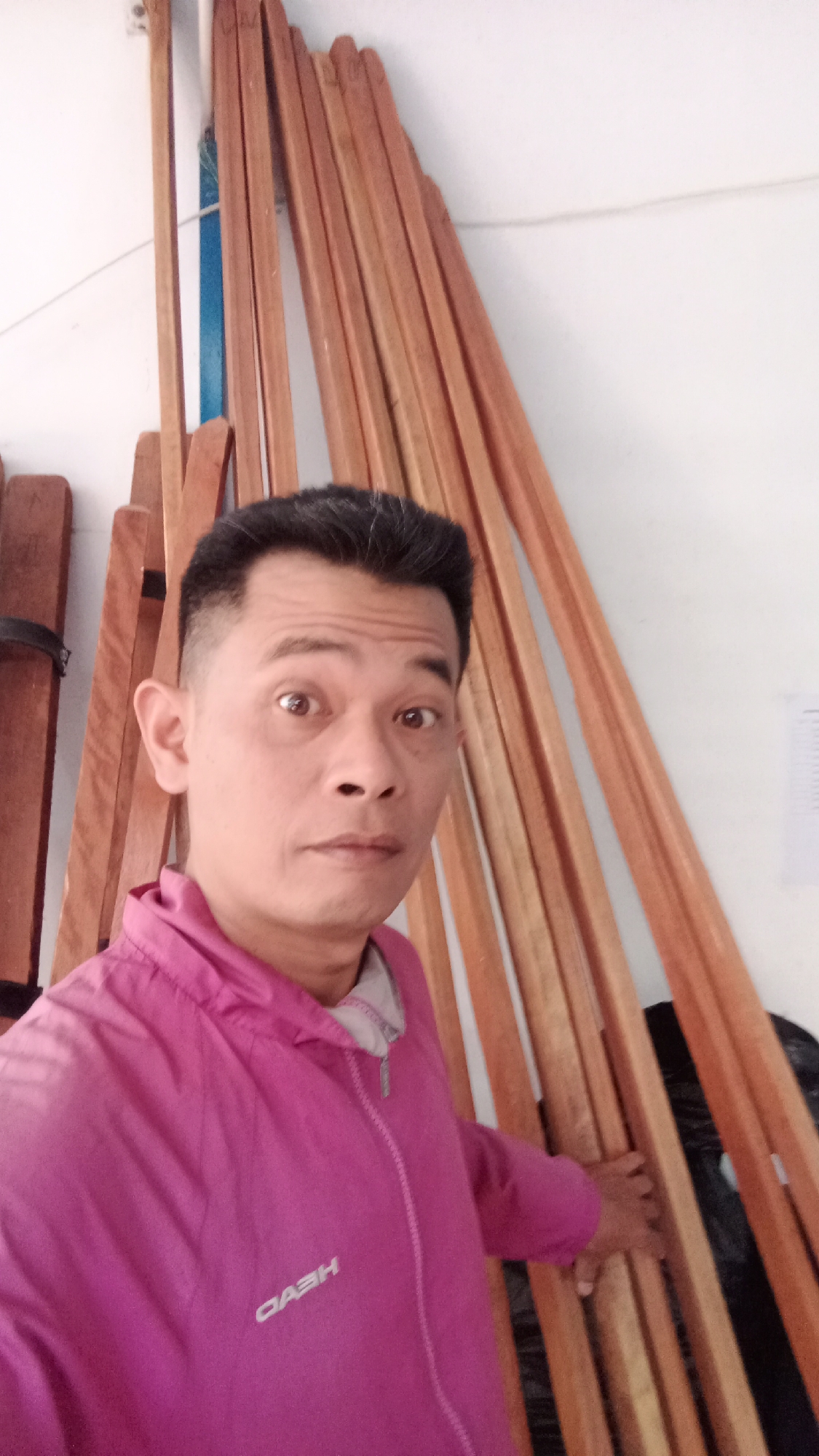 A medium-shot selfie shows a man with short, dark hair and brown eyes looking directly at the camera with a surprised expression. He is wearing a bright pink hooded jacket with a zipper, and the word "HEAD" is visible in white lettering on the front of the jacket. The man is positioned in front of a stack of long, narrow wooden planks that are leaning against a white wall. The planks are a light brown color with visible wood grain. To the left of the man, a small portion of another wooden object is visible, bound with a black strap. The lighting appears to be natural, coming from the right. The setting suggests an indoor space, possibly a workshop or storage area, located in Jawa, Indonesia.