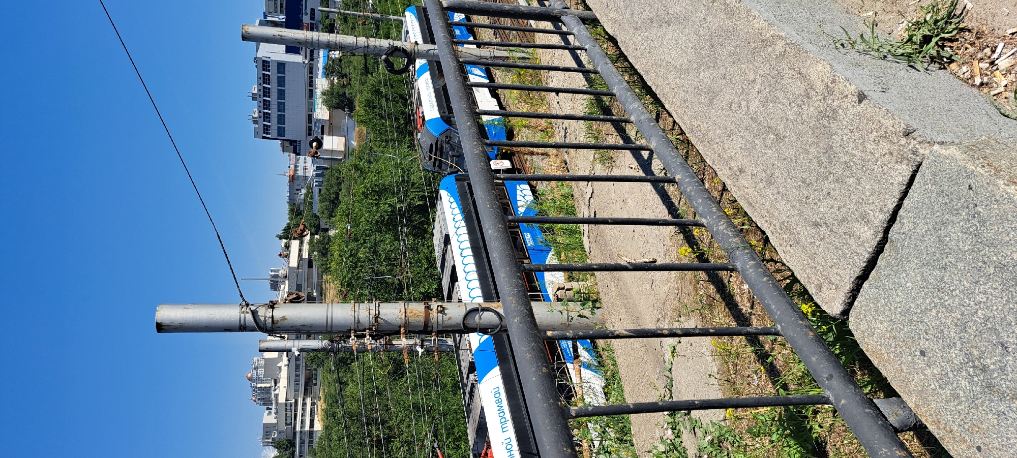 An eye-level, medium shot captures a street scene in an urban environment under a clear blue sky. The foreground is dominated by a dark, weathered metal railing, hinting at a balcony or overpass. Beyond the railing, a gray concrete barrier provides a foundational element to the scene.
Further back, a set of electric trams, adorned with striking blue and white designs, are parked on tracks. One tram has visible Russian text that reads "скоростной трамвай." The trams are situated amongst lush green trees, suggesting a park or a densely vegetated area within the city.
In the background, the cityscape unfolds with various buildings. Some are modern, multi-story structures with numerous windows, while others appear to be older constructions. The presence of utility poles and overhead wires indicates an active, functional urban setting. The overall impression is that of a sunny day in a city with well-developed public transportation.