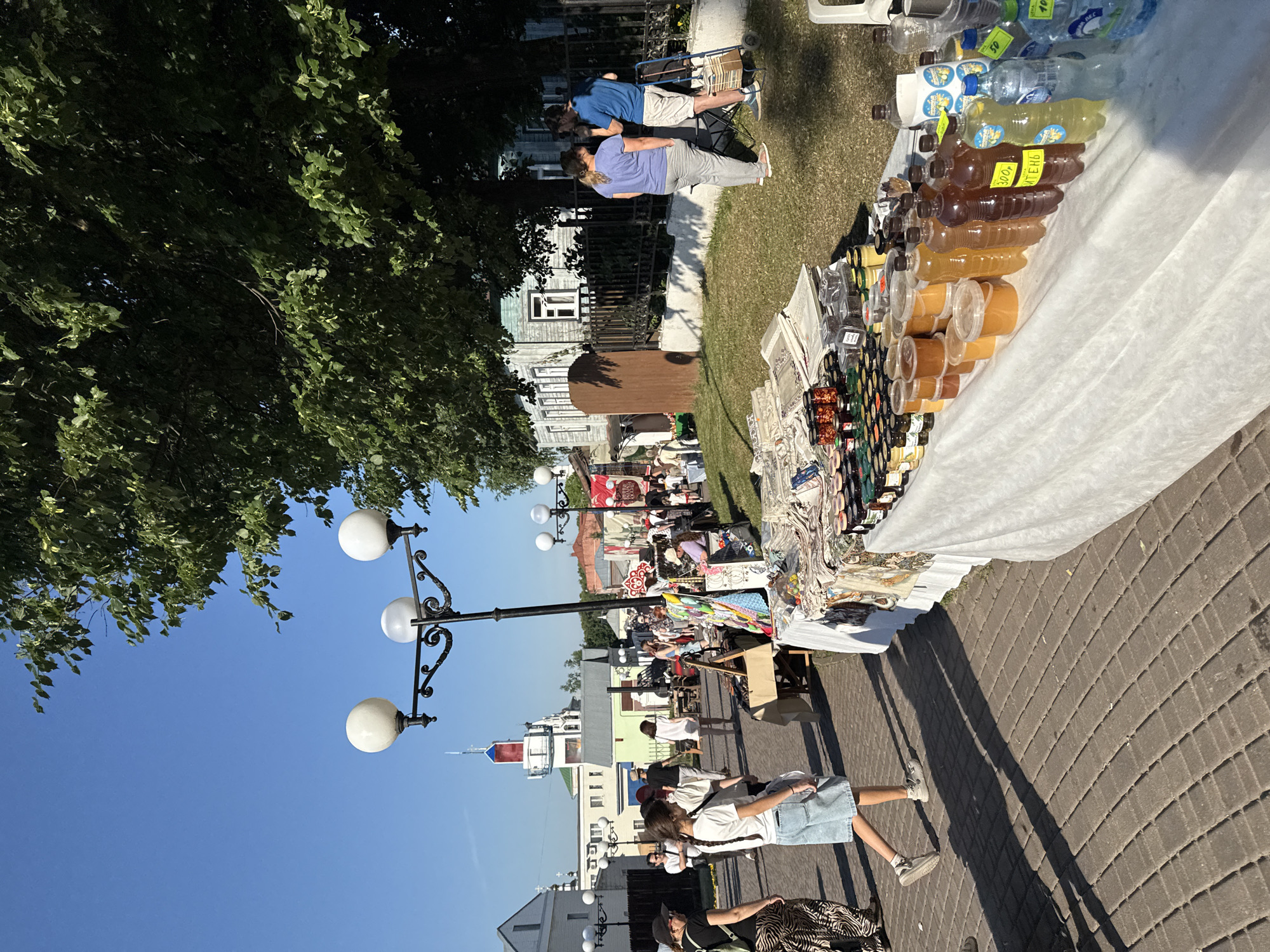 The image depicts a street market scene under a clear blue sky, likely in the city of Vladimir, Russia, given the architectural style and context of the market. In the foreground, a white-draped table displays an assortment of jars filled with honey and bottles of liquid, possibly beverages, for sale. Several people are browsing or selling at the market. To the left, a young woman wearing a white t-shirt and denim shorts walks away from the camera, her shadow cast long on the cobblestone path. Further left, other market-goers are visible, as are buildings with traditional Russian architectural elements. In the background, a distinctive white building with red accents and a tower stands out. A large tree with lush green foliage partially shades the area, casting dappled light and shadows. The overall atmosphere suggests a lively outdoor event on a sunny day.