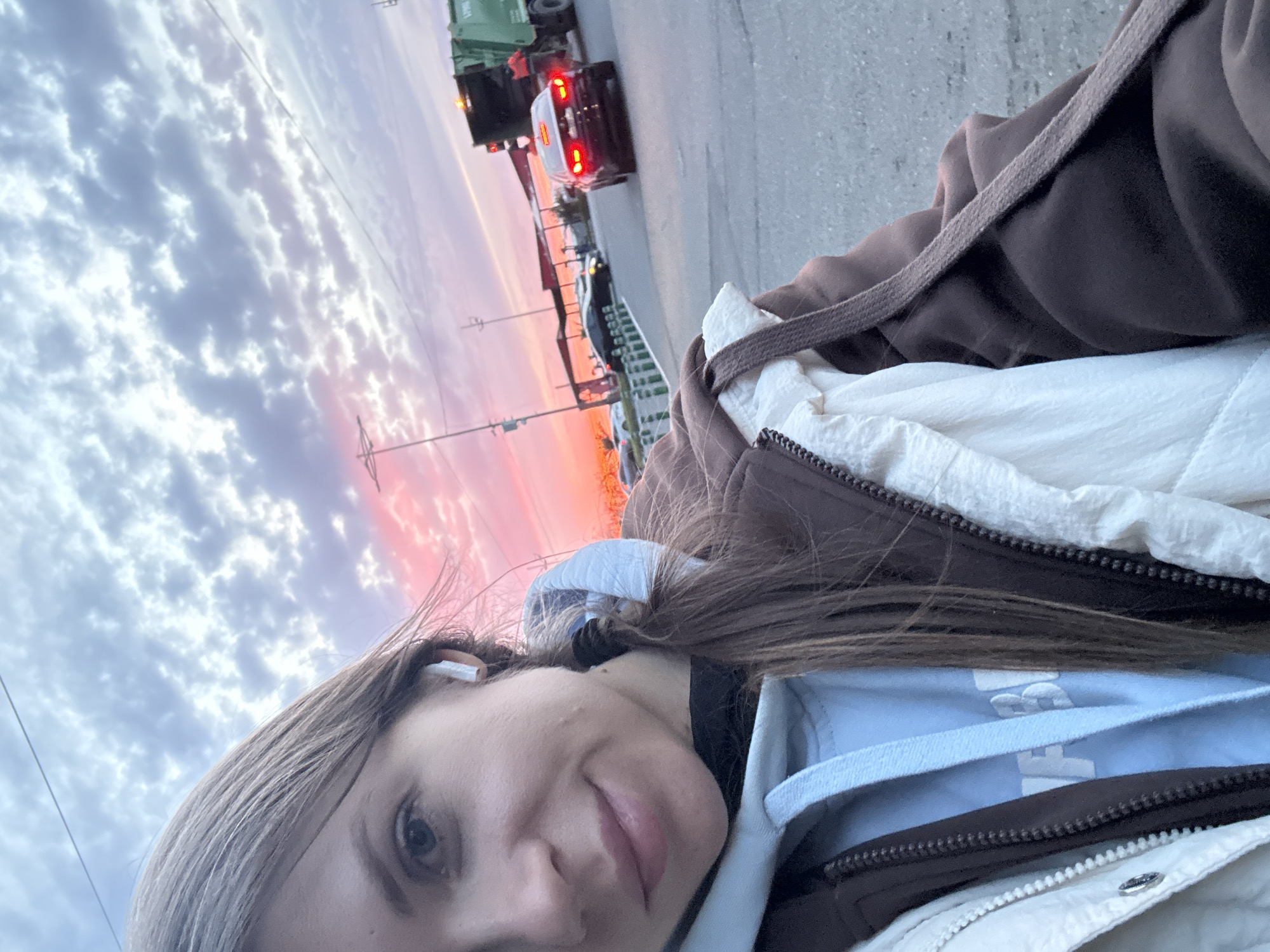 A sideways selfie shows a young woman with shoulder-length light brown hair wearing a light blue hoodie under a dark brown and white puffer jacket. An earbud is visible in her right ear. In the background, a street scene is visible during a sunset. There is a green garbage truck and a white car on the road. There is red and orange color in the sky. The city is potentially Batumi, Georgia, as per the user's request.