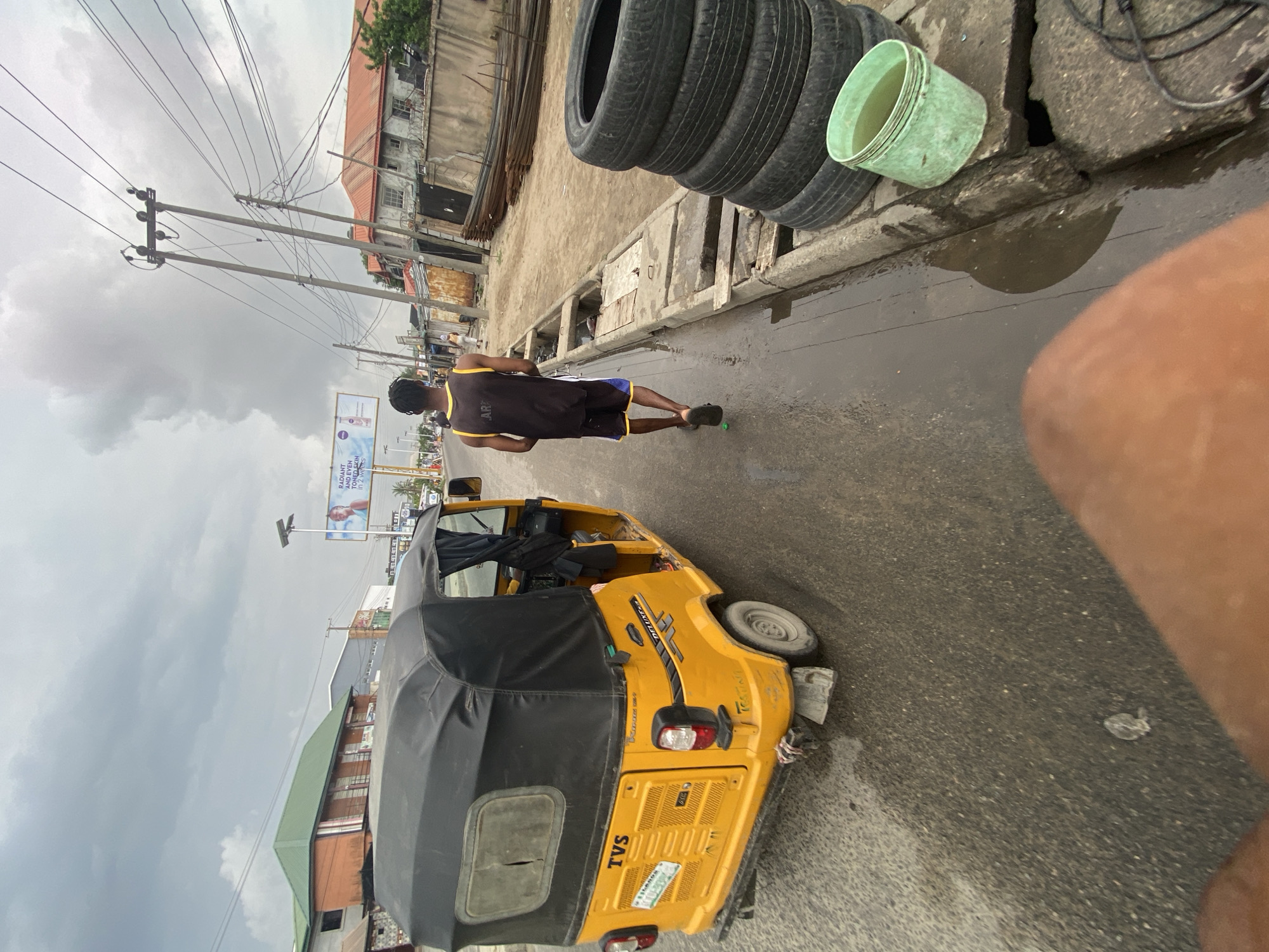 A wide-angle shot depicts a street scene on what appears to be a cloudy day in an urban setting. In the foreground, a yellow tricycle-style vehicle, possibly a "keke napep" or auto-rickshaw, sits partially on the road. A man in a dark tank top and shorts is walking away from the camera along the side of the road. Adjacent to the road is a stack of tires and a green bucket on a concrete barrier. In the background, various buildings, power lines, and a billboard are visible. The road surface appears wet, potentially indicating recent rain.