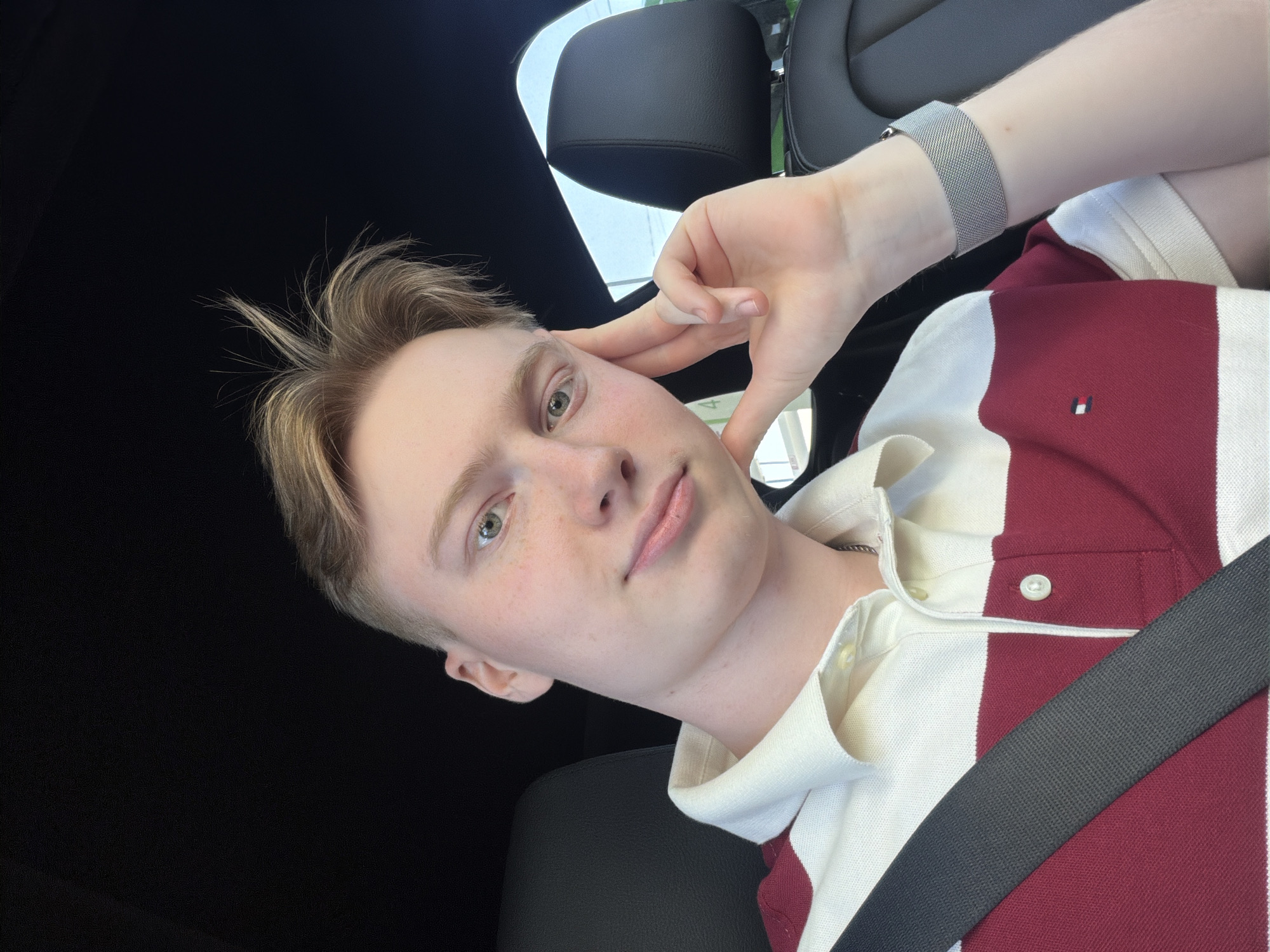A young Caucasian male with blonde hair and light green eyes is shown from the chest up, inside a car. He is wearing a red and white striped polo shirt and a silver watch on his left wrist. His right hand is resting on his cheek, and his head is tilted slightly. The background shows the dark interior of a car, including a black seat and a sliver of a window with a light blue sky visible. There is no indication of the city of Kyiv, Ukraine, or any other specific location within the image.
